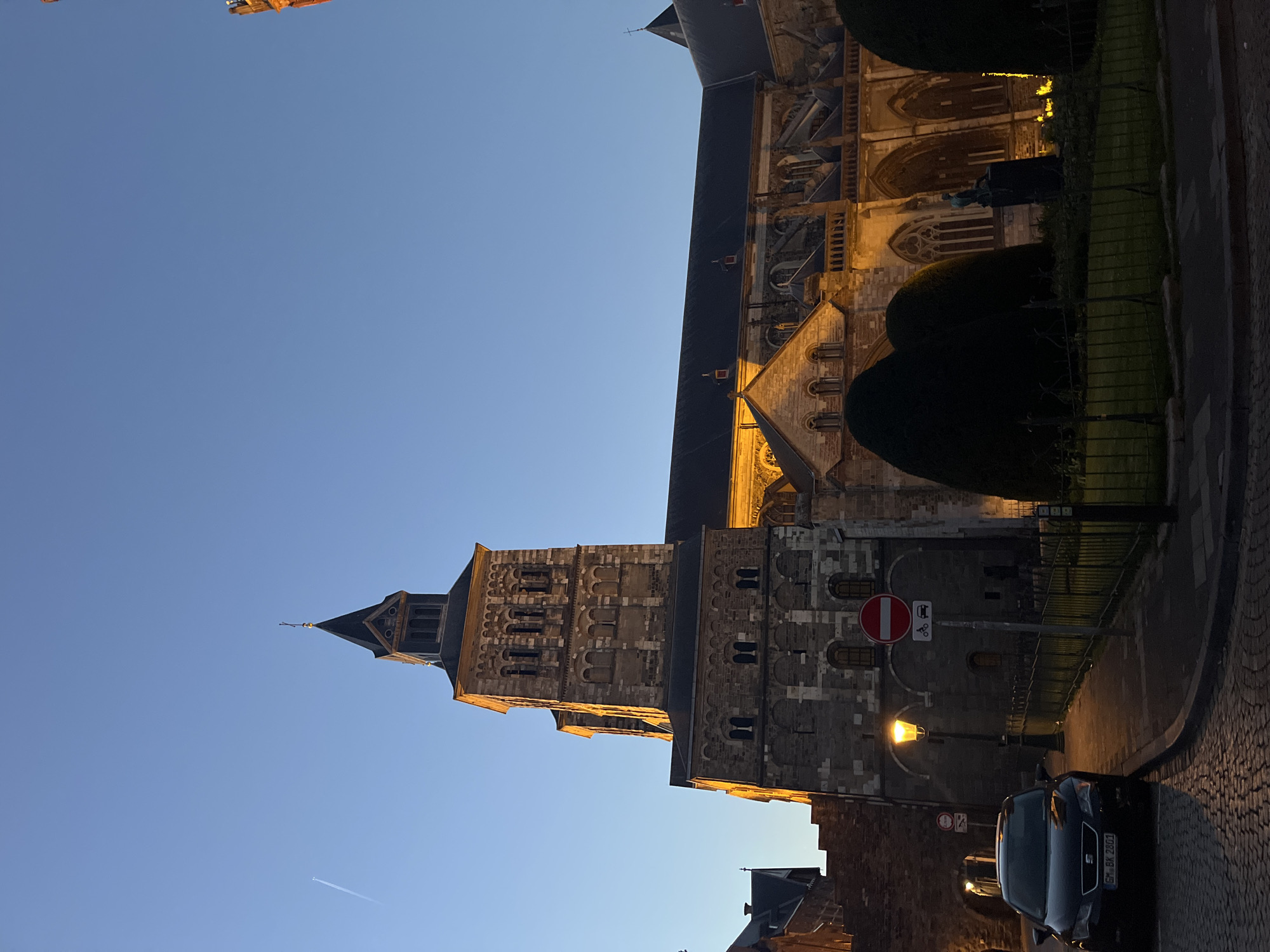 The image depicts the Basilica of Saint Servatius in Maastricht, Netherlands, from a low-angle street-level perspective at dusk. The sky is a clear gradient of blue. The basilica's stone tower rises prominently, illuminated by artificial lights that highlight its architectural details. A portion of the basilica's roofline and facade are visible. Street-level elements include a street lamp, a road sign indicating "no entry", trimmed bushes, a paved street, and a parked dark-colored car. A contrail is seen in the sky.