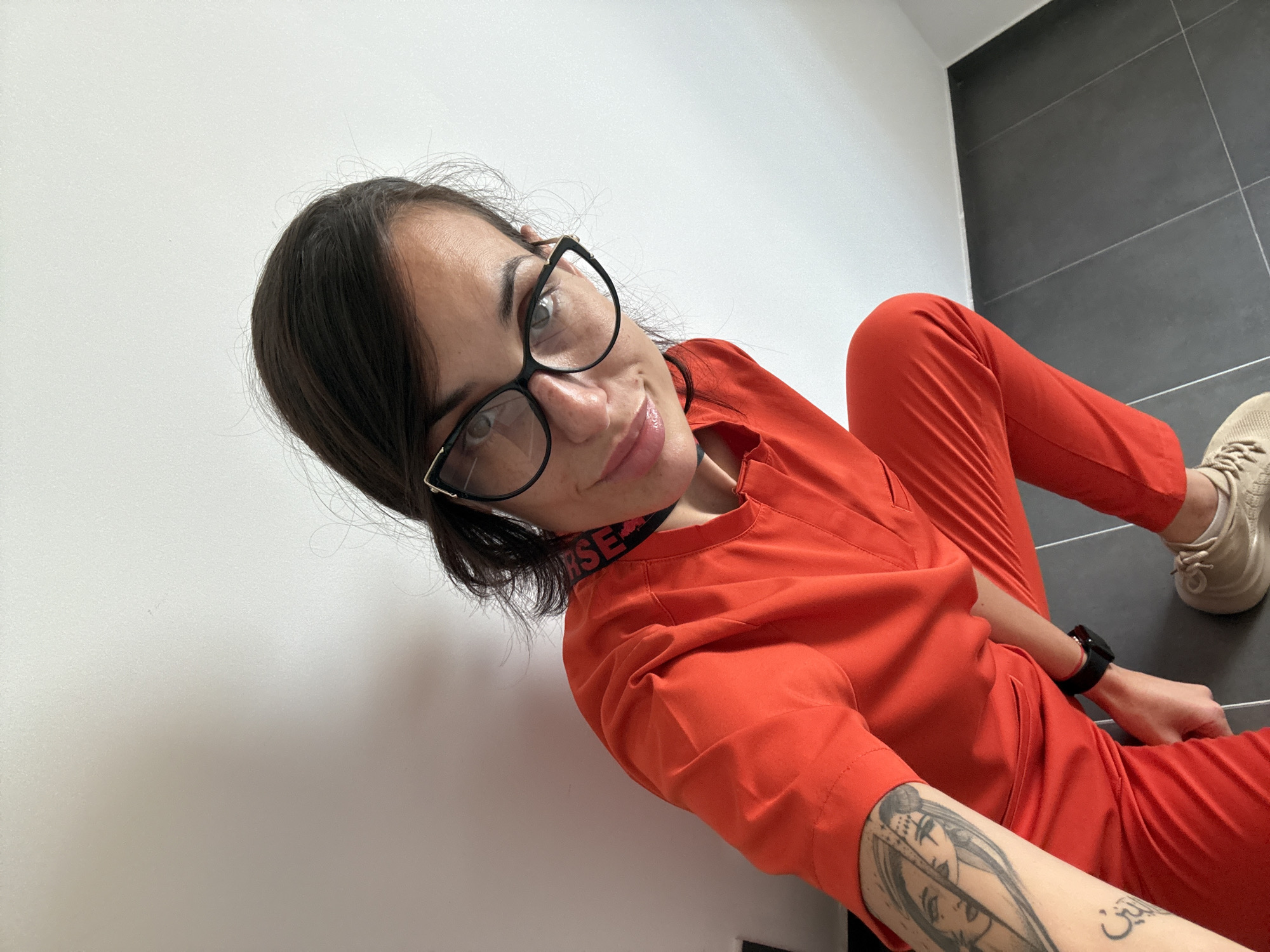 A young woman with dark hair, wearing eyeglasses and orange scrubs with "NURSE" visible on the neckline, is lying on the floor. She has a tattoo on her left forearm and wears a watch on her left wrist. Her beige sneakers are visible on the right side of the frame. The floor consists of gray tiles. The background includes a white wall. This scene could hypothetically occur in a medical facility in Pleven, Bulgaria, but there is no explicit evidence for this conclusion from the image.