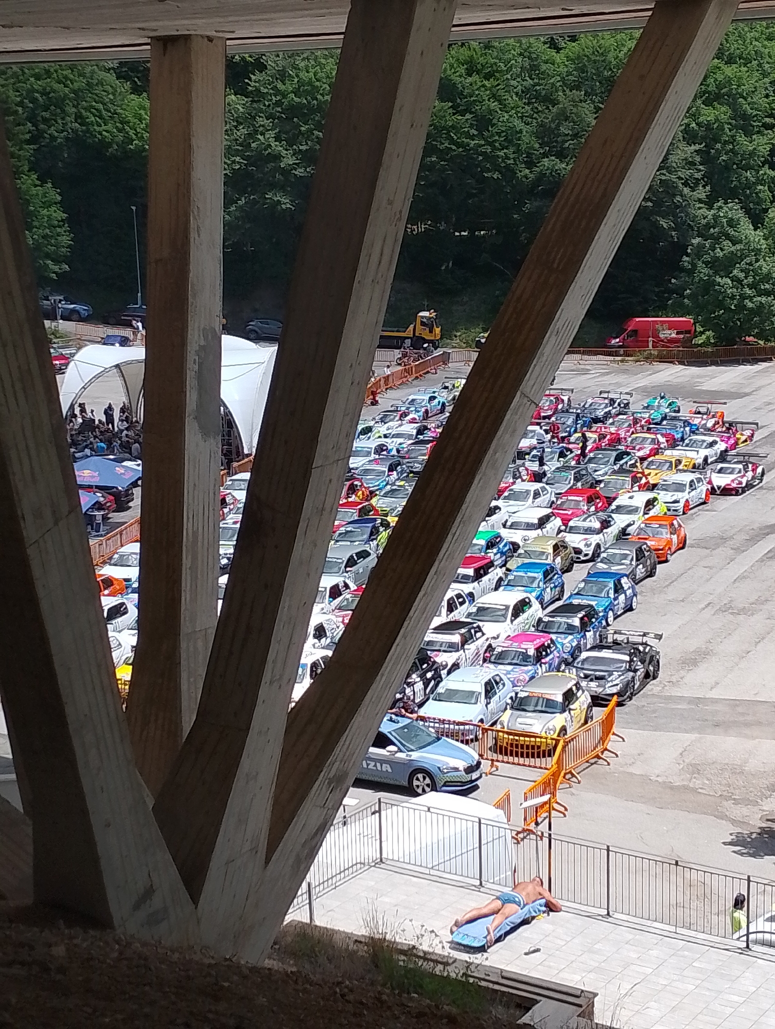 The image depicts an outdoor event, possibly a race or car show, set in a large paved area. In the foreground, large concrete pillars from a bridge or overpass dominate the view, casting shadows and framing the scene. Beyond the pillars, a multitude of colorful cars, many of them appear to be modified or racing vehicles with spoilers and racing decals, are parked in neat rows. Some of the cars are white, red, blue, yellow, and black, with various sponsor logos and designs.
There are also several people visible. A person is lying on their back on a blue mat on a tiled area, with a black mobile phone or remote beside them. Further away, near the cars, several individuals are standing and observing the vehicles. There's also a police car with "POLIZIA" written on it. Tents or canopies are set up in the background, suggesting a service or spectator area. The environment is characterized by a bright, sunny day, with lush green trees forming a backdrop behind the event area.
The caption specifies the location as the city of Rieti, Italy. Given the landscape and the presence of what appears to be a racing event or car gathering, this scene could be taking place at a venue like the Autodromo di Rieti or a similar open area within or on the outskirts of the city. The overpass structure in the foreground is a distinctive architectural element.