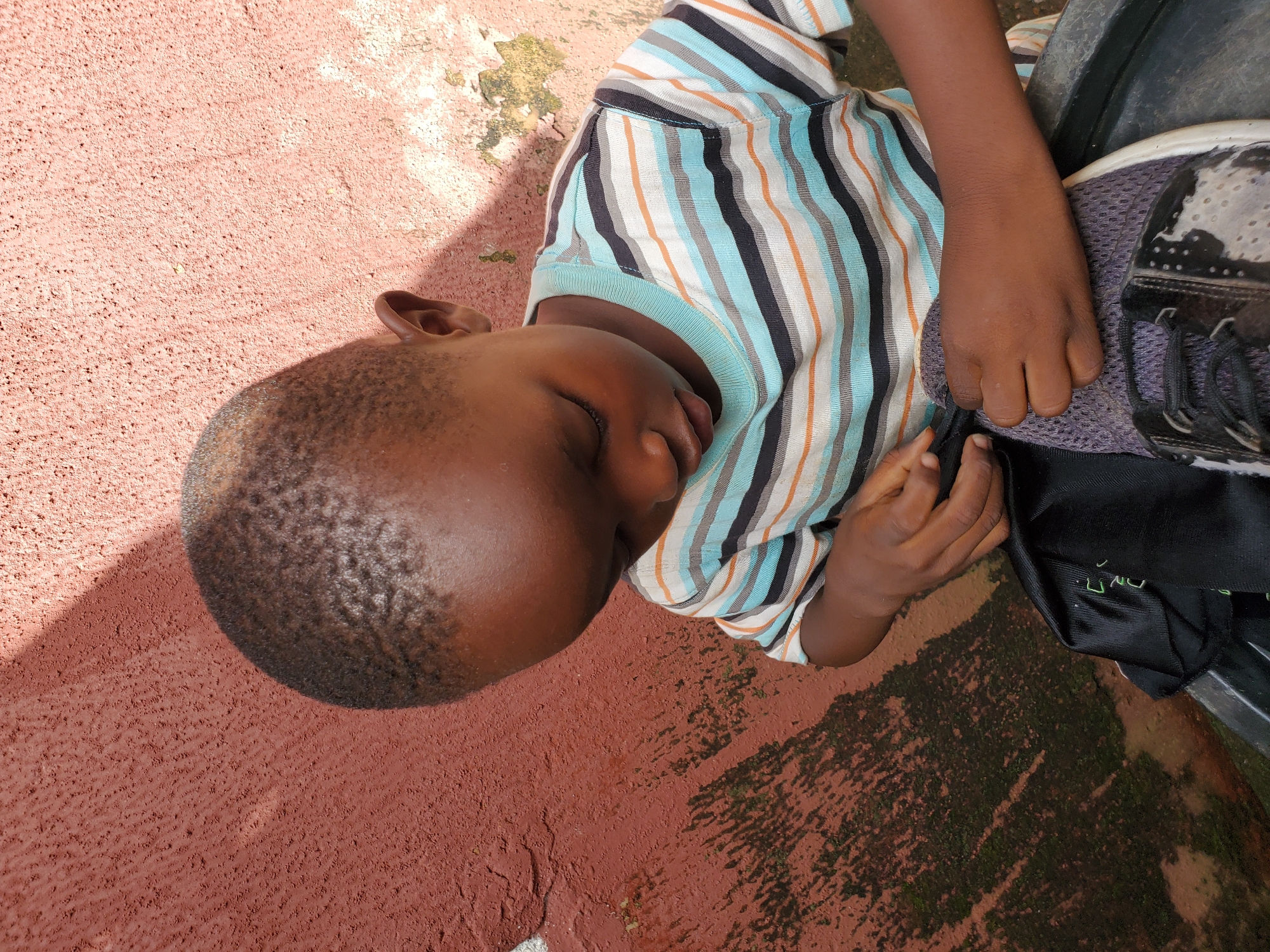 This media file shows a young child lying down on a textured, reddish-brown surface, possibly pavement or ground. The child is wearing a striped t-shirt with bands of light blue, white, orange, and black. Their eyes are closed, and their head is turned to the side. The child's left arm is resting on their chest, and their right hand is near their face. A shoe with black and white detailing and black laces is visible next to the child. The scene is outdoors, with shadows indicating sunlight. Some patches of moss or algae are visible on the ground. The setting appears to be an urban or semi-urban area in Jos, Nigeria. No text is visible in the image.