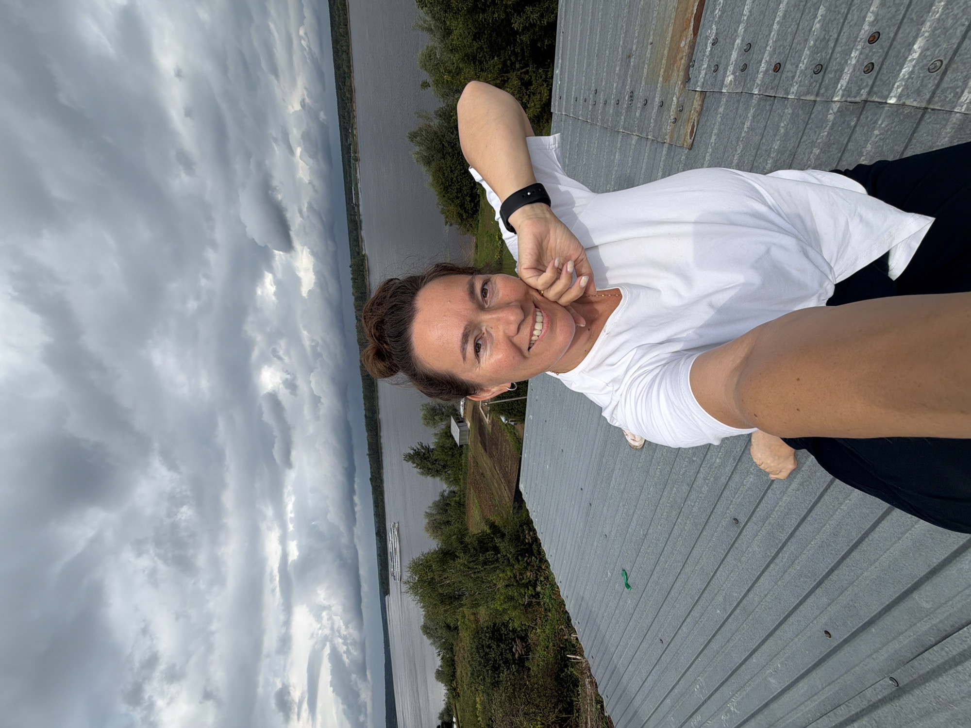 A woman is taking a selfie on a corrugated metal roof in Olkhovka, Russia. She has her hair up in a bun and is wearing a white t-shirt and black pants. Her left arm is bent at the elbow and resting on her head, and her right arm is extended, holding the camera. She is smiling at the camera. In the background, a wide river is visible, with trees and fields on the opposite bank. A large white ship is sailing on the river. The sky is cloudy and gray.