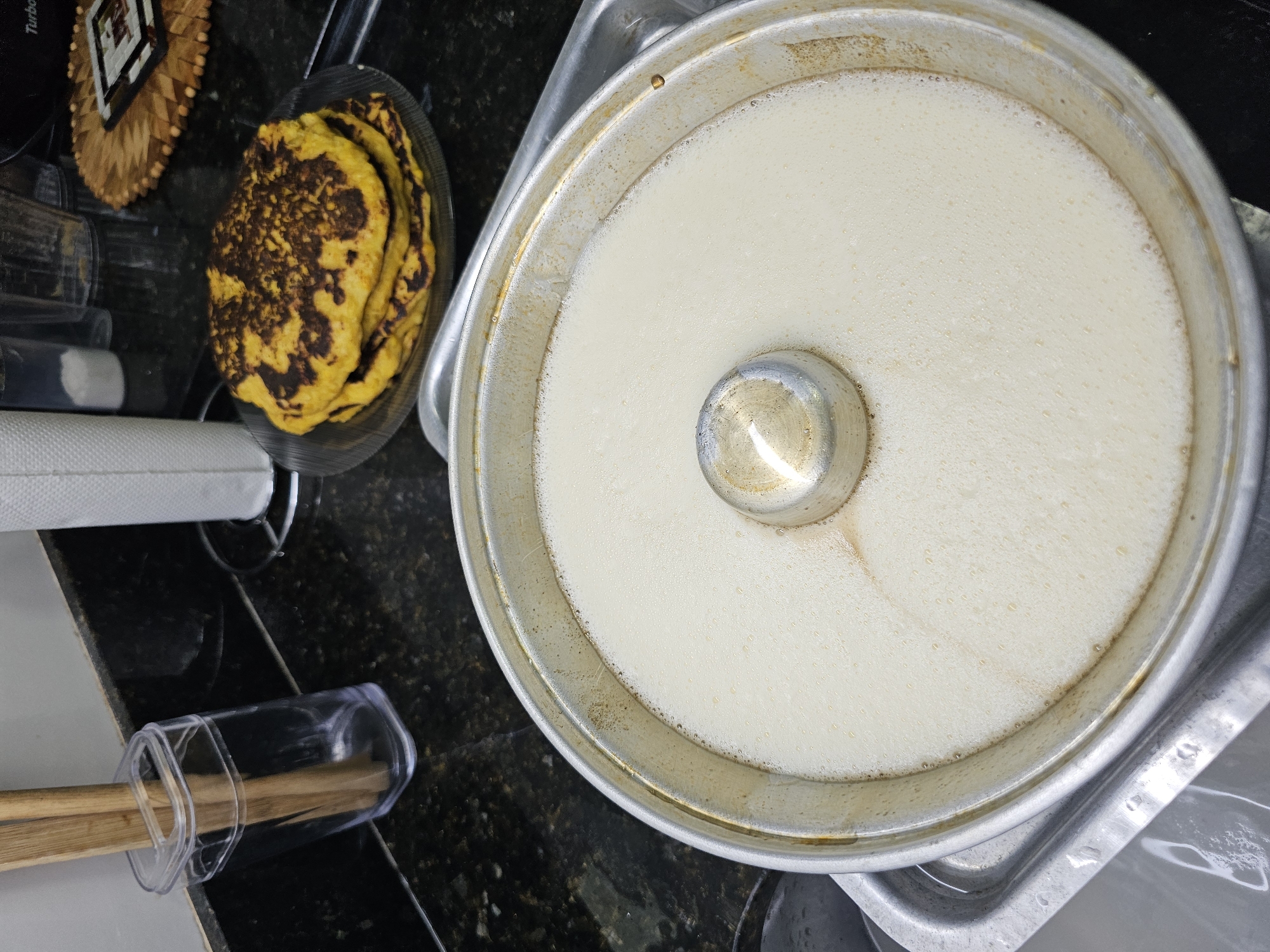 The image shows a metal bundt cake pan filled with a light-colored liquid, likely a batter or custard. The pan sits in a larger metal tray. To the left of the pan, there is a plate containing several yellow pancakes or similar items with dark spots, stacked on top of each other. A wooden placemat, a roll of paper towels, and utensils in a clear container are also visible on the left side of the image. The background appears to be a kitchen counter with a dark, speckled surface. The location and city are unknown.