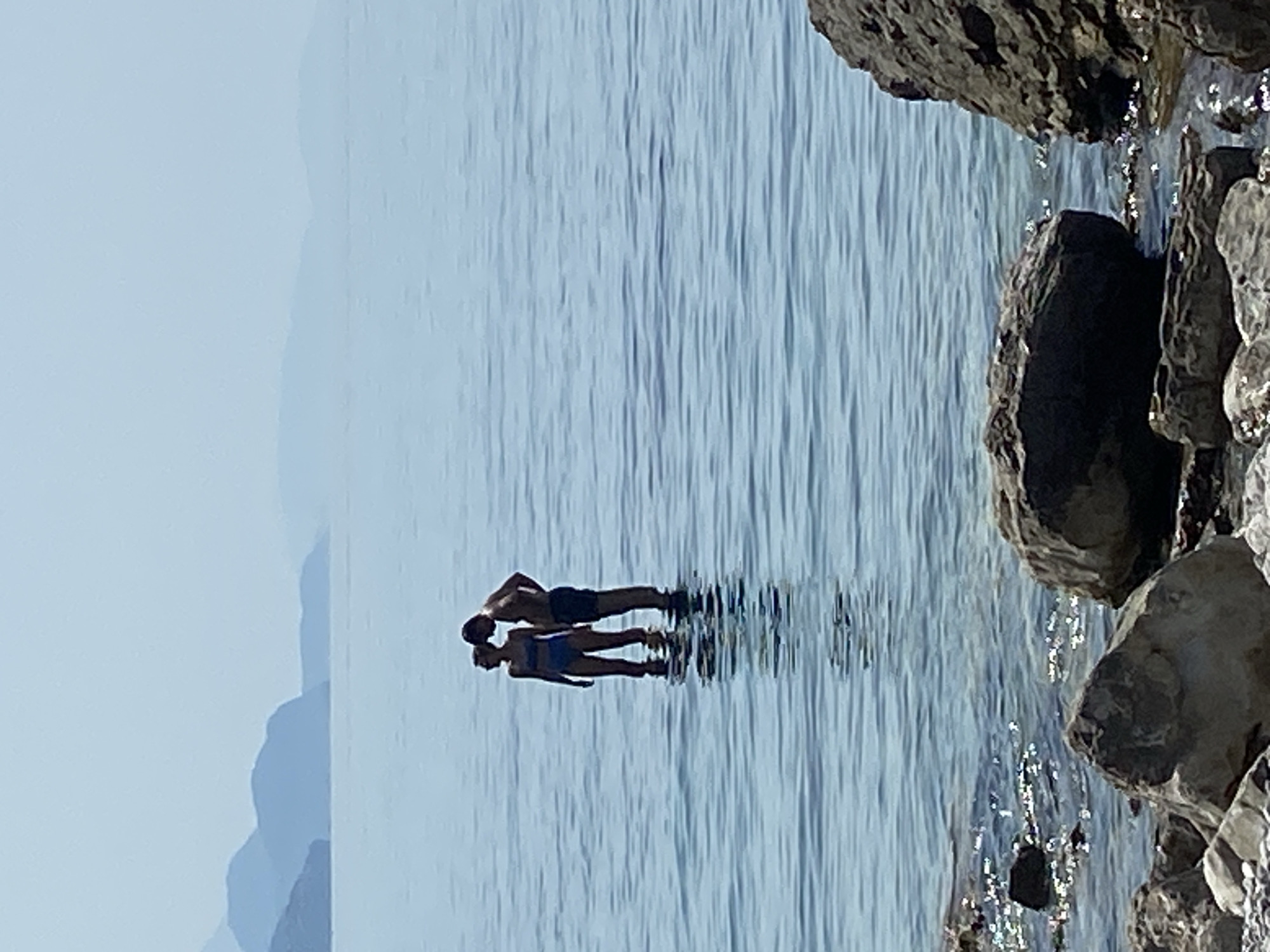 Two individuals, a male and a female, stand in shallow, clear blue water. The female, wearing a blue bikini, faces left and slightly away from the male. The male, wearing dark swim trunks, faces the female, with his head tilted down as if speaking to her. They appear to be in a serene, natural environment with a hazy mountainous landscape in the background, visible across a vast expanse of calm water. The foreground is occupied by a rocky shoreline. The scene suggests a peaceful moment shared between two people on a body of water, possibly a bay or the sea, in the vicinity of Prijevor, Montenegro.