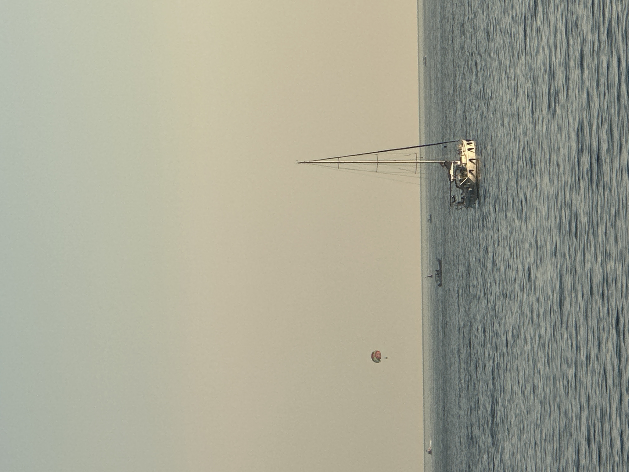 A sailboat is docked in the calm waters off the coast of Budva, Montenegro. The sailboat has a white hull with dark trim and a tall mast with sails that are furled. The sky is a pale, hazy blue, and the sun is low in the sky, casting a warm glow over the scene. In the distance, a person is parasailing, tethered to a boat. Several other small boats are also visible on the water. The overall impression is one of a peaceful and serene day at sea.