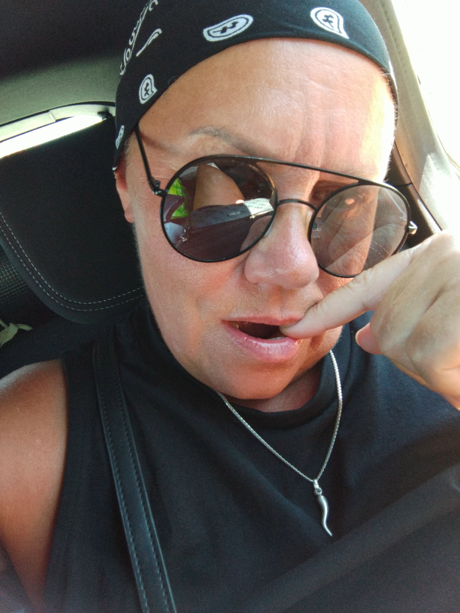A close-up, selfie-style photograph captures a person sitting in the driver's seat of a car, likely in Vieste, Italy, given the context. The individual, wearing a black bandana with white paisley patterns and dark sunglasses, is looking at the camera with their right index finger touching their lips. They are also wearing a black top and a silver necklace with a horn-shaped pendant. The background shows the interior of a car, including a headrest and part of the dashboard. The lighting suggests it is daytime, possibly sunny. The person's facial expression is ambiguous, but the finger-to-lip gesture could imply thoughtfulness or shyness. The overall impression is a candid moment captured during a drive.