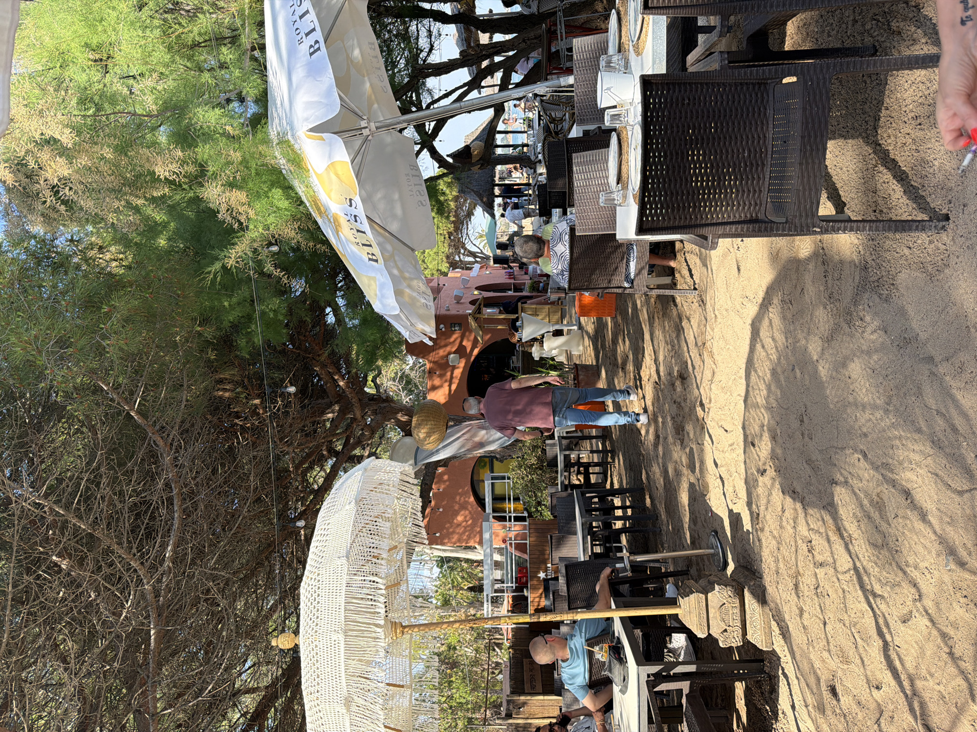 The image shows an outdoor dining area located on the beach in Mont-roig del Camp, Spain. Several tables and chairs are arranged on a sandy surface, indicating the beach setting. Some tables are set for dining, with plates and glasses visible. People are present, seated and walking. Parasols provide shade over some of the tables. Trees and vegetation surround the dining area. The architecture of nearby buildings is visible in the background.