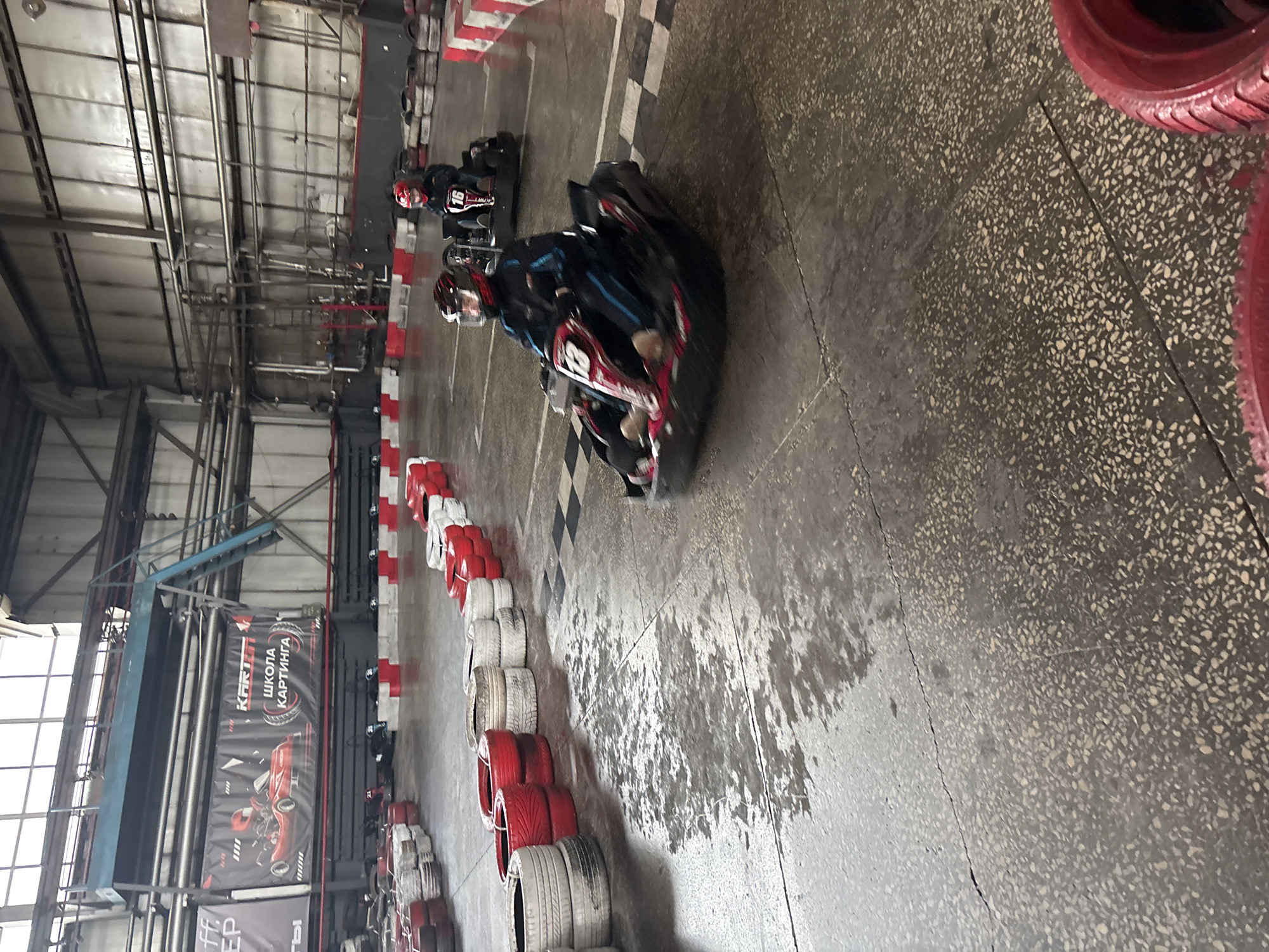 A medium-shot photograph captures a go-karting race in progress inside a large indoor facility, possibly located in Perm, Russia given the Cyrillic signage in the background. Two go-karts are visible, with the lead kart closer to the viewer, driven by a person wearing a helmet and dark clothing. The kart is turning sharply, and the driver is leaning into the turn. Another kart, with the number "16" visible, follows behind. The go-kart track is surrounded by a barrier of stacked tires painted in red and white. The floor is concrete. The building's interior is industrial, with metal beams and pipes visible overhead. A banner in Cyrillic hangs from the rafters. There is ample ambient lighting.