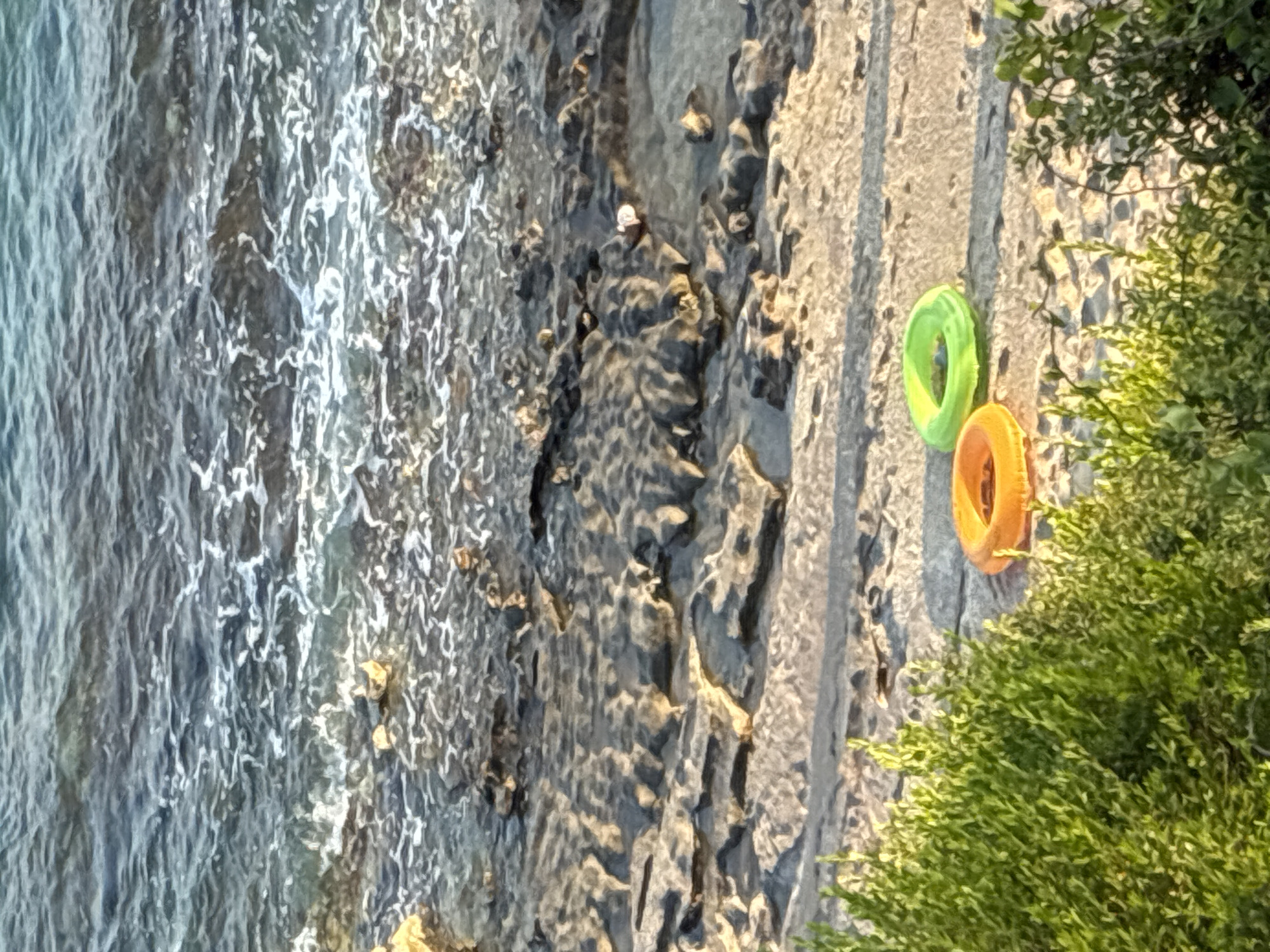 The image shows a rocky coastline in Alanya, Turkey, with the sea in the background. Two inflatable rings, one orange and one green, lie on a flat rocky surface near the water's edge. Lush green vegetation borders the lower portion of the scene. The tide is coming in, with waves washing over the rocks. The lighting suggests it is either early morning or late afternoon, casting long shadows on the rocky terrain.