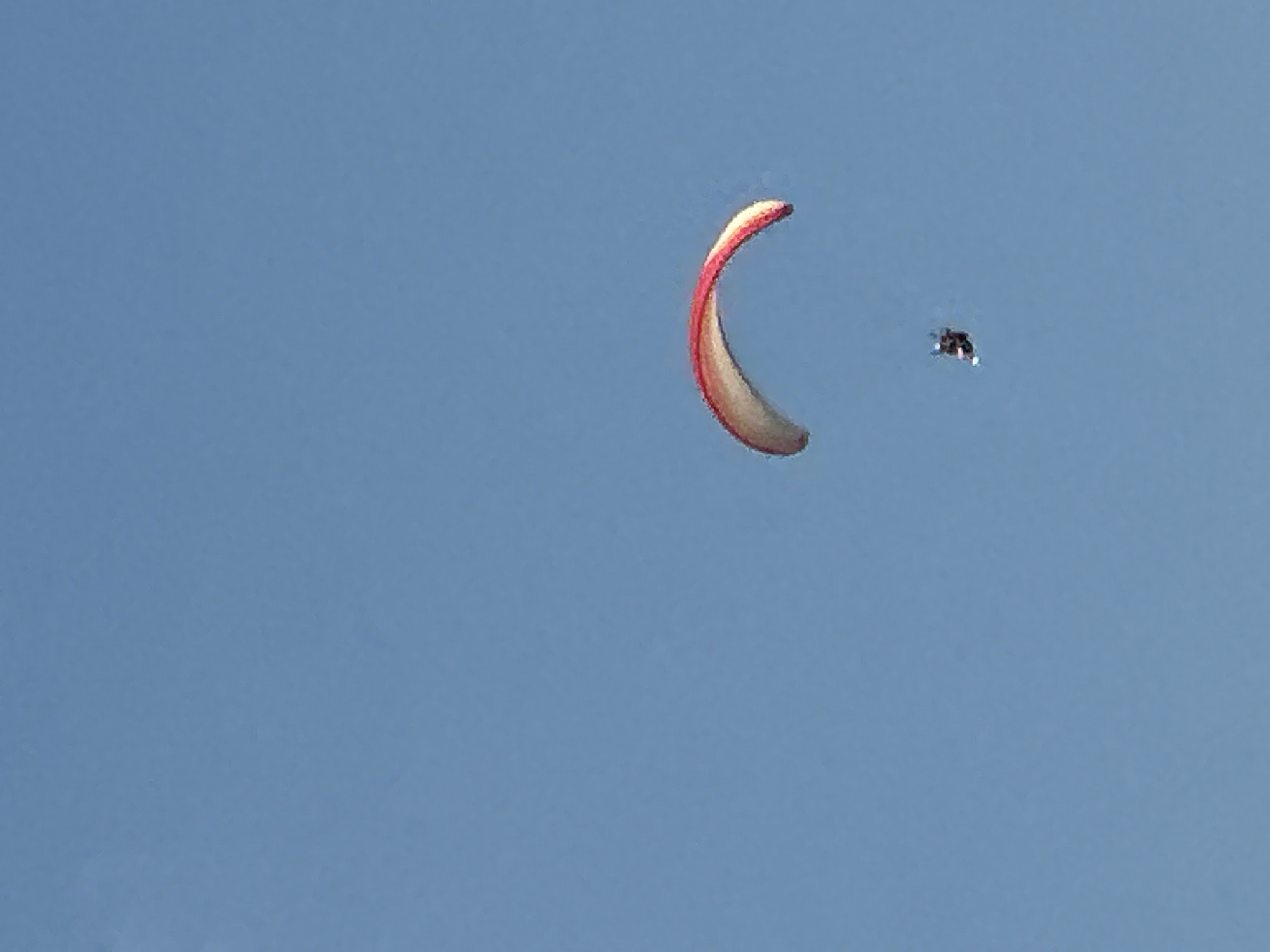 The image shows a paraglider soaring through a clear blue sky. The paraglider, a red and white wing, is curved and open, suspended above a small figure in a dark suit. The paraglider is positioned in the mid-right of the frame, with the figure directly below it. The background is an uninterrupted expanse of blue sky, indicating an outdoor activity. The image does not provide any specific information about the location in the city of Boreti, Montenegro, as no landmarks or identifying features are visible.