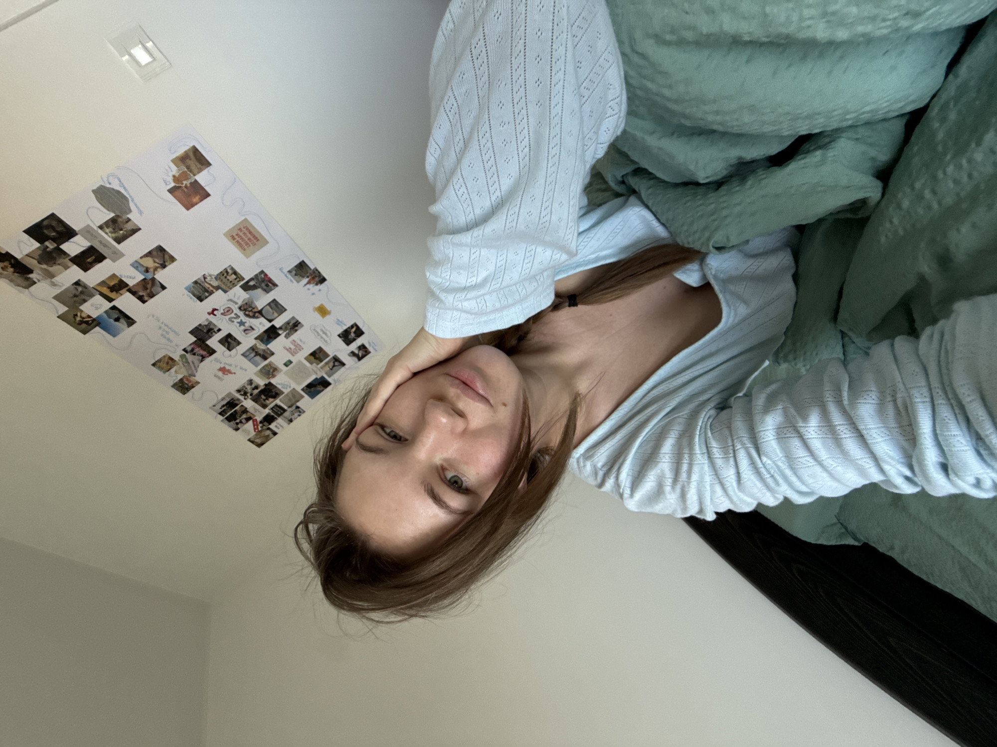 The scene depicts an individual inside a room in what is identified as the city of Vozdovac Urban Municipality, Serbia.
**Subject:** A light-skinned female individual with light brown, slightly disheveled hair, partially tied into a braid on her right side, is positioned in the foreground, facing the camera directly. Her eyes are green/hazel. She exhibits a natural complexion with visible small moles or freckles on her face. She is wearing a light blue, long-sleeved top featuring an intricate patterned knit texture. Her left hand is placed against her left cheek/jawline.
**Actions and Interactions:** The individual is engaged in a self-portrait, looking directly into the camera with a neutral expression, indicating she is aware of being photographed. She appears to be reclining or lying down, partially covered by bedding.
**Environment and Objects:**
* **Bed:** A dark headboard, possibly wood, is visible on the left side of the frame.
* **Bedding:** The individual is covered by a crumpled, textured, mint green duvet or blanket.
* **Wall:** The background consists of a plain white wall.
* **Wall Decorations:** On the upper right portion of the wall, a large white sheet or board is affixed. This board is decorated with numerous small, square photographs or printed images, arranged in a collage. Blue lines and handwritten text, including "2+2=6" and some Cyrillic script, are present on the board.
* **Wall Fixture:** To the right of the collage, a white, rectangular electrical fixture, likely a light switch or outlet cover, is mounted on the wall.
**Lighting:** The room is illuminated by what appears to be natural light, resulting in soft shadows and a clear view of the scene.