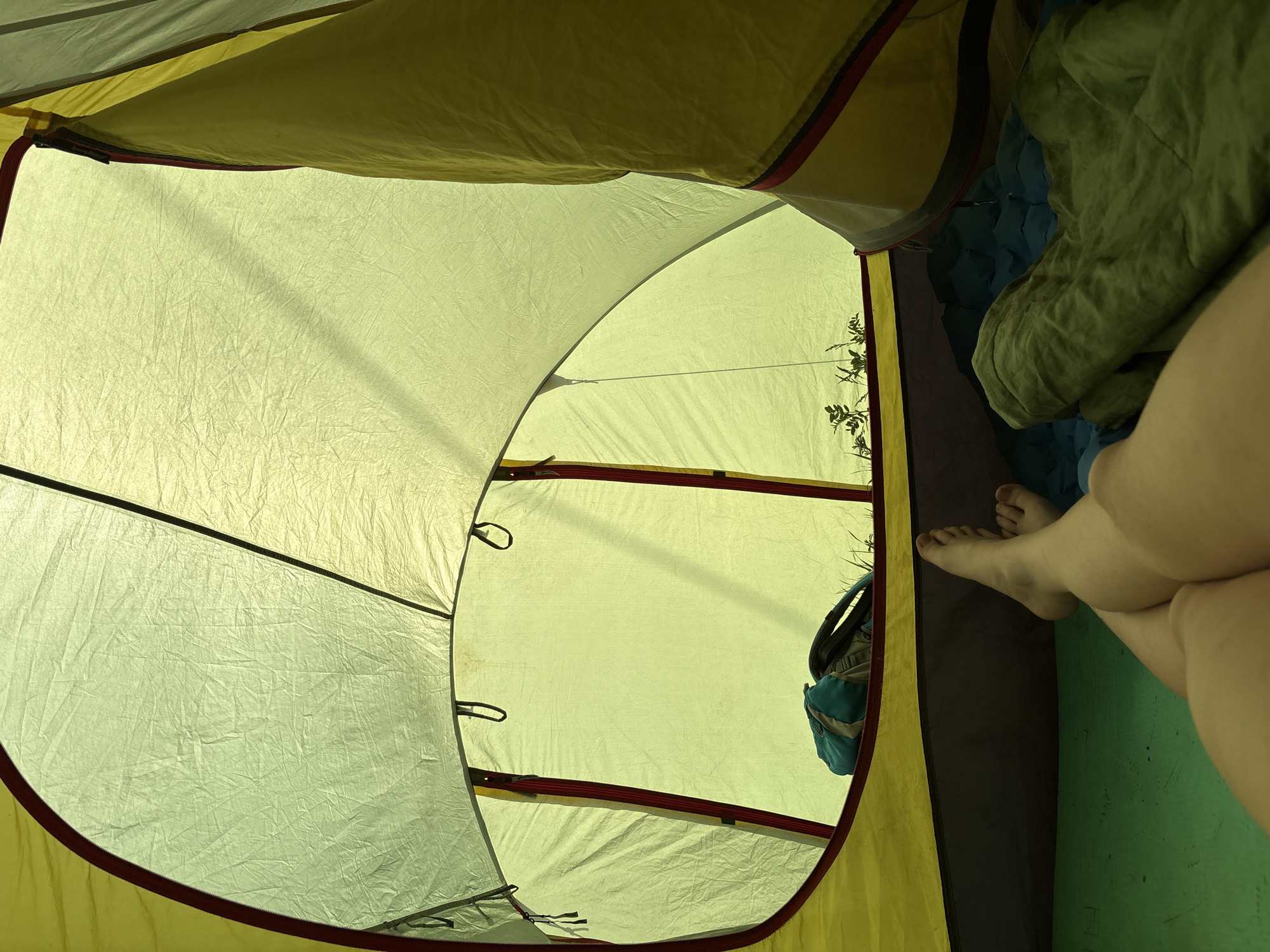 A low-angle, interior view of a tent reveals a person's bare feet and legs in the right of the frame, suggesting they are lying down. Next to the legs is a green sleeping bag on top of a blue sleeping pad. The interior of the tent is made of a yellow/beige material. In the doorway of the tent, a patch of green vegetation is visible, along with a backpack. The location is unknown and cannot be derived from the image.