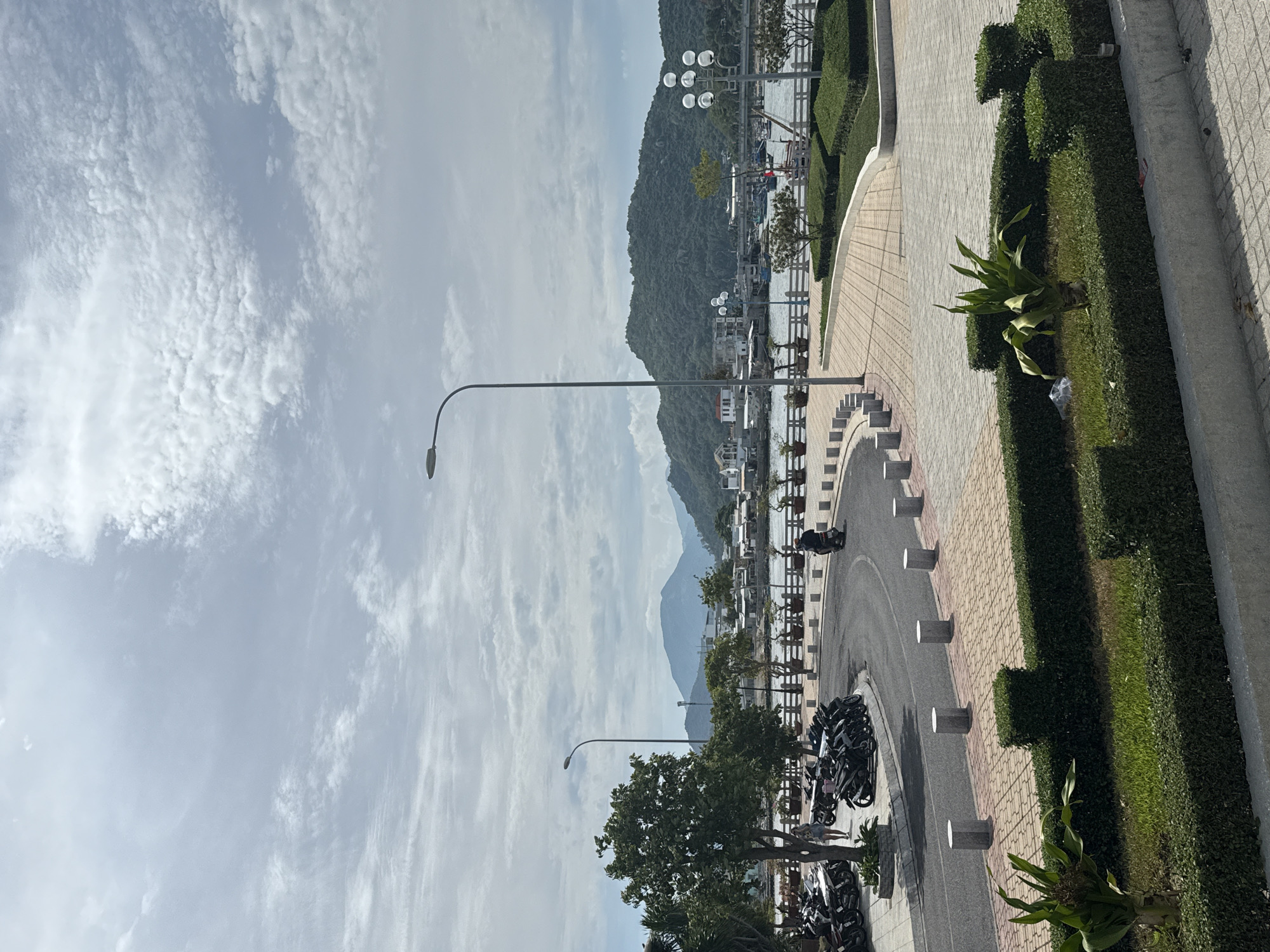 The image captures a scene in Nha Trang Ward, Vietnam. The view is from an elevated position, looking towards a body of water, possibly a bay or river, with a large, green mountain in the background. The sky is mostly cloudy.
In the foreground and middle ground, there's a paved road curving around, bordered by a tiled walkway and a row of closely-trimmed hedges and decorative plants. A series of cylindrical concrete bollards lines the edge of the road and walkway. Several parked motorcycles are visible along the roadside.
Tall streetlights are positioned along the road. In the distance, structures and boats are visible on the water near the base of the mountain, implying a harbor or residential area. One person is discernible among the parked motorcycles.