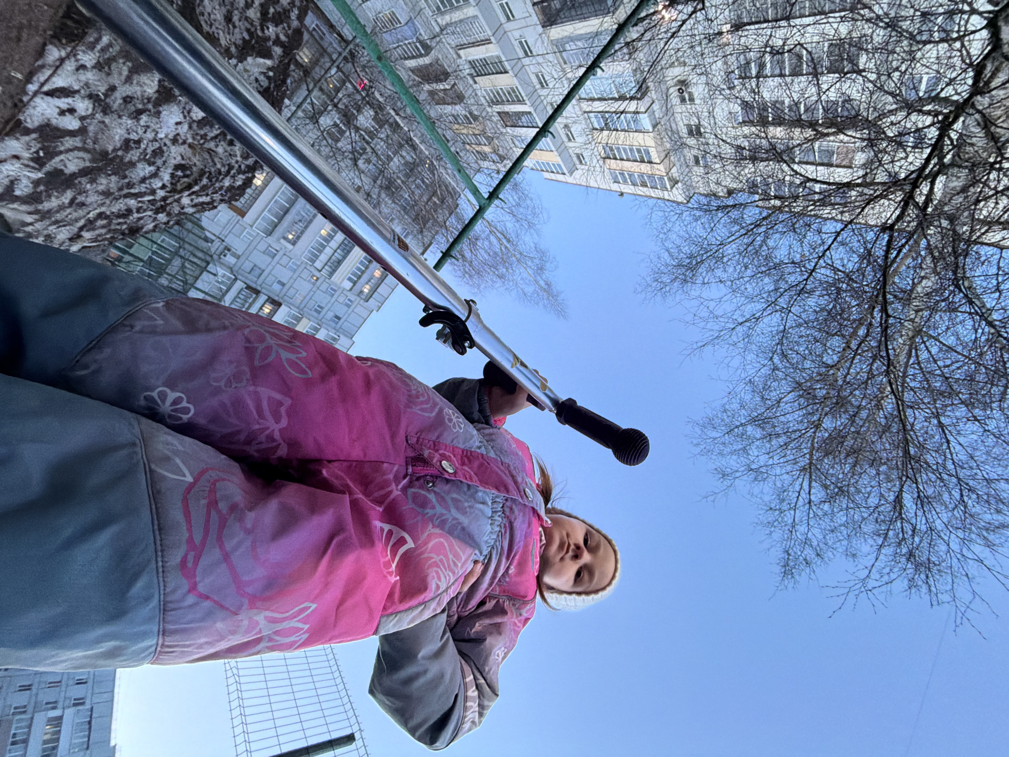 The image, taken from a low-angle perspective looking upwards, features a young child holding a scooter handlebar. The child is wearing a pink, purple, and gray patterned winter jacket or snowsuit and a white knitted hat. They are looking directly at the camera with a neutral expression. The scooter has a prominent silver metal T-bar handle.
In the background, against a clear bright blue sky, are several large, multi-story residential buildings with many windows, suggesting an urban or suburban setting. Bare trees, indicative of winter or early spring, are visible throughout the upper portion of the frame, especially on the right side. To the far left, there's a patch of ground covered with what appears to be dirty snow or ice. A green metal fence or railing is also partially visible at the bottom left.
The activity shows the child standing with their scooter, likely posing for the photograph. The time of day is daytime, under a bright, cloudless sky. The presence of bare trees and snow/ice points to cold weather conditions. No visible text is present in the image. The scene is consistent with an urban outdoor environment in a location like Chaikovsky, Russia, during winter or early spring.