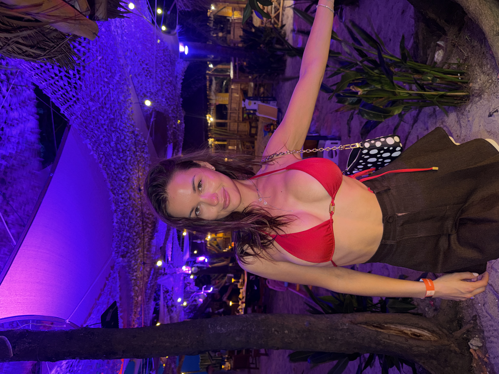 A woman is sitting in a restaurant, posing for a picture. She is wearing a red bikini top and brown high-waisted pants. She has a black and white polka dot purse over her shoulder. The restaurant is located in Choeng Thale, Thailand. The restaurant is decorated with lights and plants.