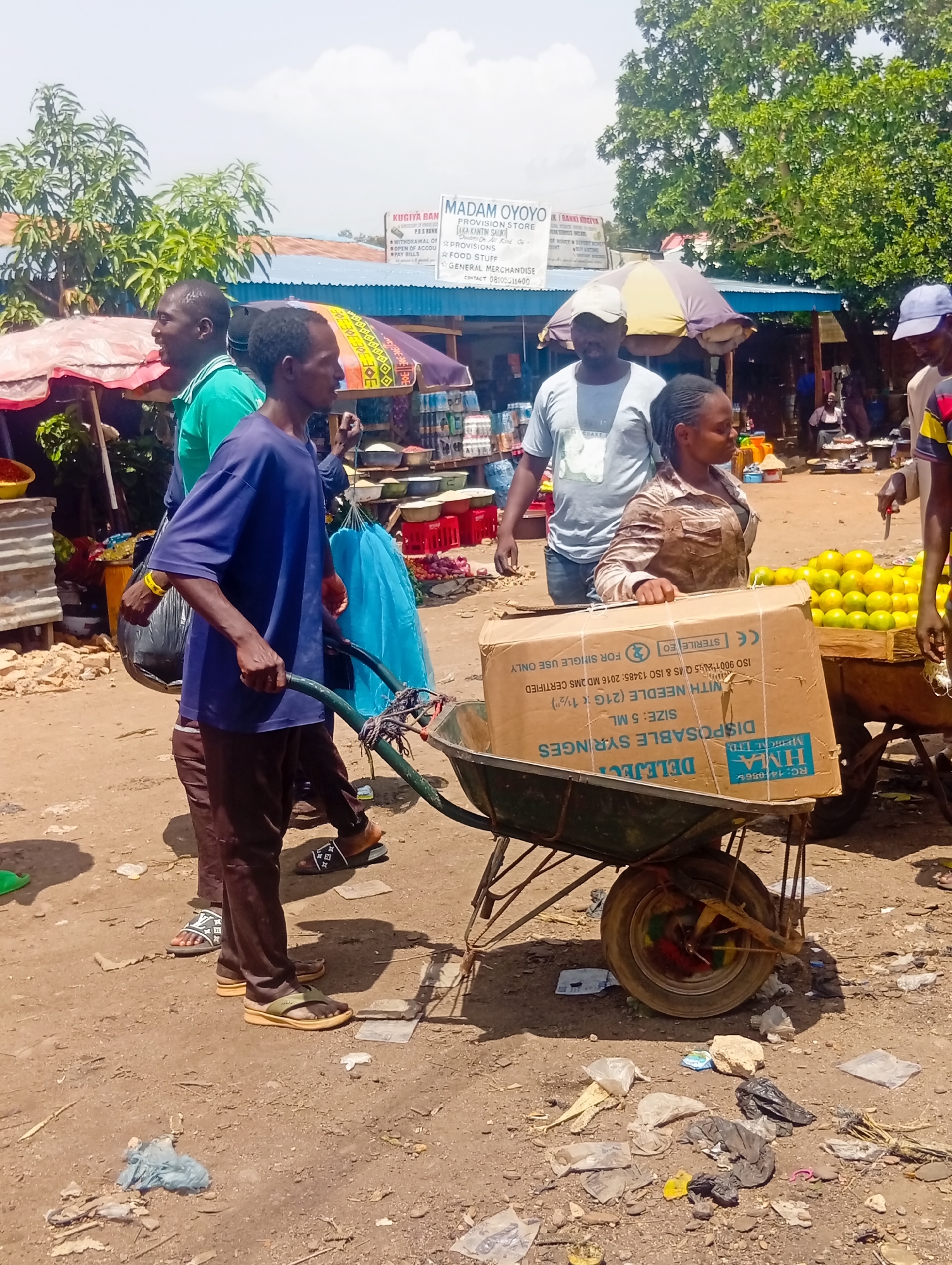 In a bustling outdoor market scene in Bukuru, Nigeria, three men and one woman are captured amidst the activity. The foreground features a man in a blue t-shirt and brown trousers pushing a green wheelbarrow loaded with a large cardboard box. The box is labeled with English text, including "DISPOSABLE SYRINGES" and "SIZE: 5 ML." To his left, another man in a green t-shirt and brown trousers walks beside him, carrying a black plastic bag. Behind them, a woman in a light brown patterned shirt and a man wearing a grey t-shirt and a white cap are visible. In the background, market stalls with colorful awnings line the dusty ground. Piles of oranges are displayed on a wooden cart to the right, and a variety of goods, including what appear to be household items and produce, are arranged at the stalls. The environment is open-air with trees and structures suggesting a typical Nigerian marketplace.
