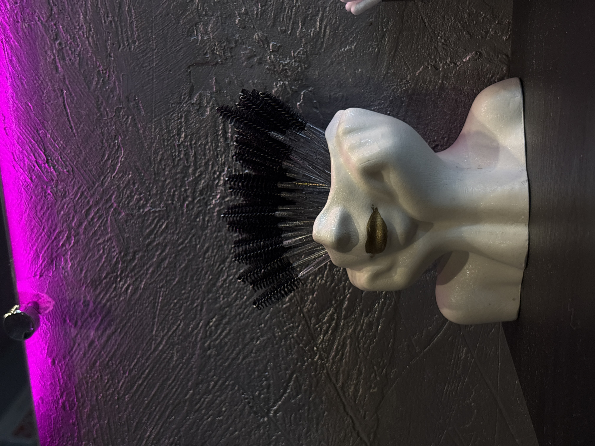 A close-up, low-angle shot features a white bust planter, shaped like a stylized human face with its hands propping up its head, filled with black spoolie brushes. The planter has gold-colored lips and rests on a dark surface. Behind the planter, a textured, dark gray wall is illuminated by a bright pink light source positioned at the top of the frame. A small, reflective object is visible within the pink light. The scene is indoors, likely in a salon or beauty-related establishment in Rostov-on-Don, Russia.