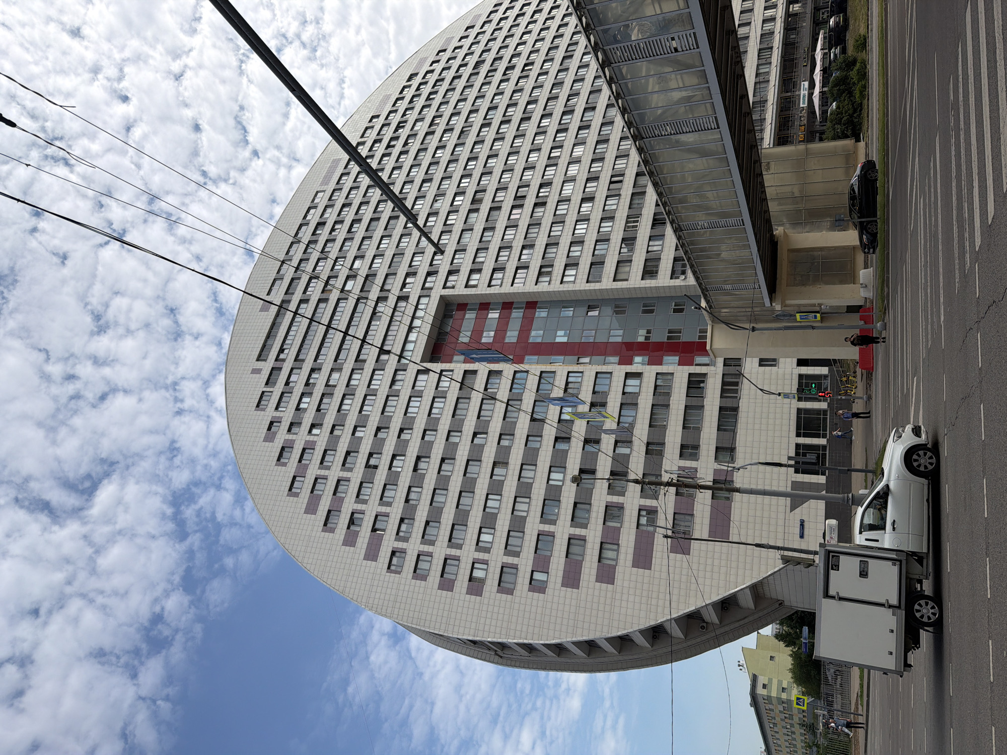 This image depicts the building known as "House of the Book" or "Dom Knigi" located in Moscow, Russia. The distinct crescent-shaped, multi-story building dominates the frame, its facade clad in light-colored tiles with dark and purple accents around the numerous windows. A covered pedestrian bridge, enclosed in glass, extends across the foreground, connecting to the building.
In the street below, a white refrigerated truck is driving on the asphalt road. A black sedan is parked on the side of the road near the pedestrian bridge. Several pedestrians are visible on the sidewalks and crossing the street at a marked crosswalk. The sky above is partly cloudy with a mix of white cumulus clouds against a blue sky. Electrical wires are strung across the upper portion of the image. The environment appears to be an urban setting with a mix of modern architecture.
