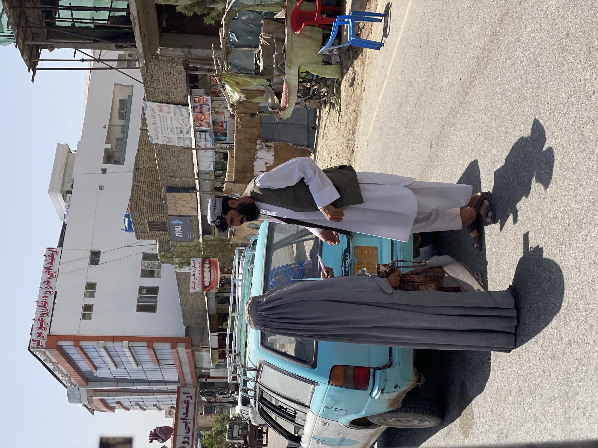 The image depicts a street scene in Kandahar, Afghanistan. In the foreground, a man in traditional clothing, including a white shalwar kameez and a dark vest, stands facing a person covered in a grey burqa, who is standing by the open rear door of a light blue station wagon. The man is holding a small, light-colored object in his outstretched hand, seemingly interacting with the person in the burqa. The station wagon appears old and weathered, with a roof rack and visible "COROLLA" badging on the rear.
In the background, a modern multi-story building with glass windows and orange accents dominates the scene. Signs with writing in Arabic script are visible on the building and on smaller structures to the right. One sign features an advertisement for dental services with images of teeth. A street vendor's stall, covered with a fabric awning and containing various items and seating, is also present. The environment suggests a sunny day, with clear skies and distinct shadows cast by the subjects and objects. The road appears to be asphalt.