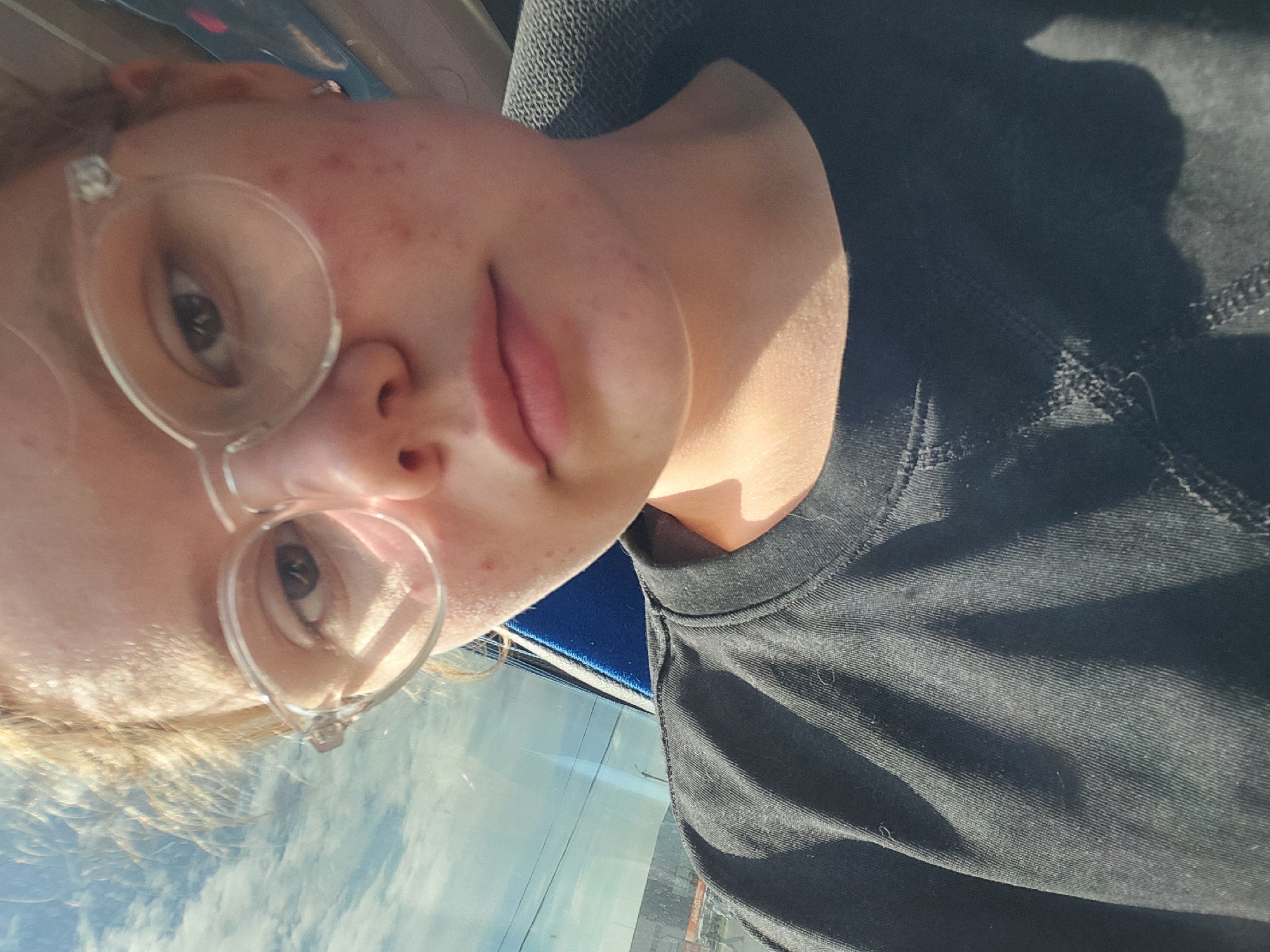 A selfie of a person with fair skin, wearing clear-framed glasses and a black shirt. The person is likely on public transportation with a window showing buildings and clouds in the background. The location is Unknown, Unknown.