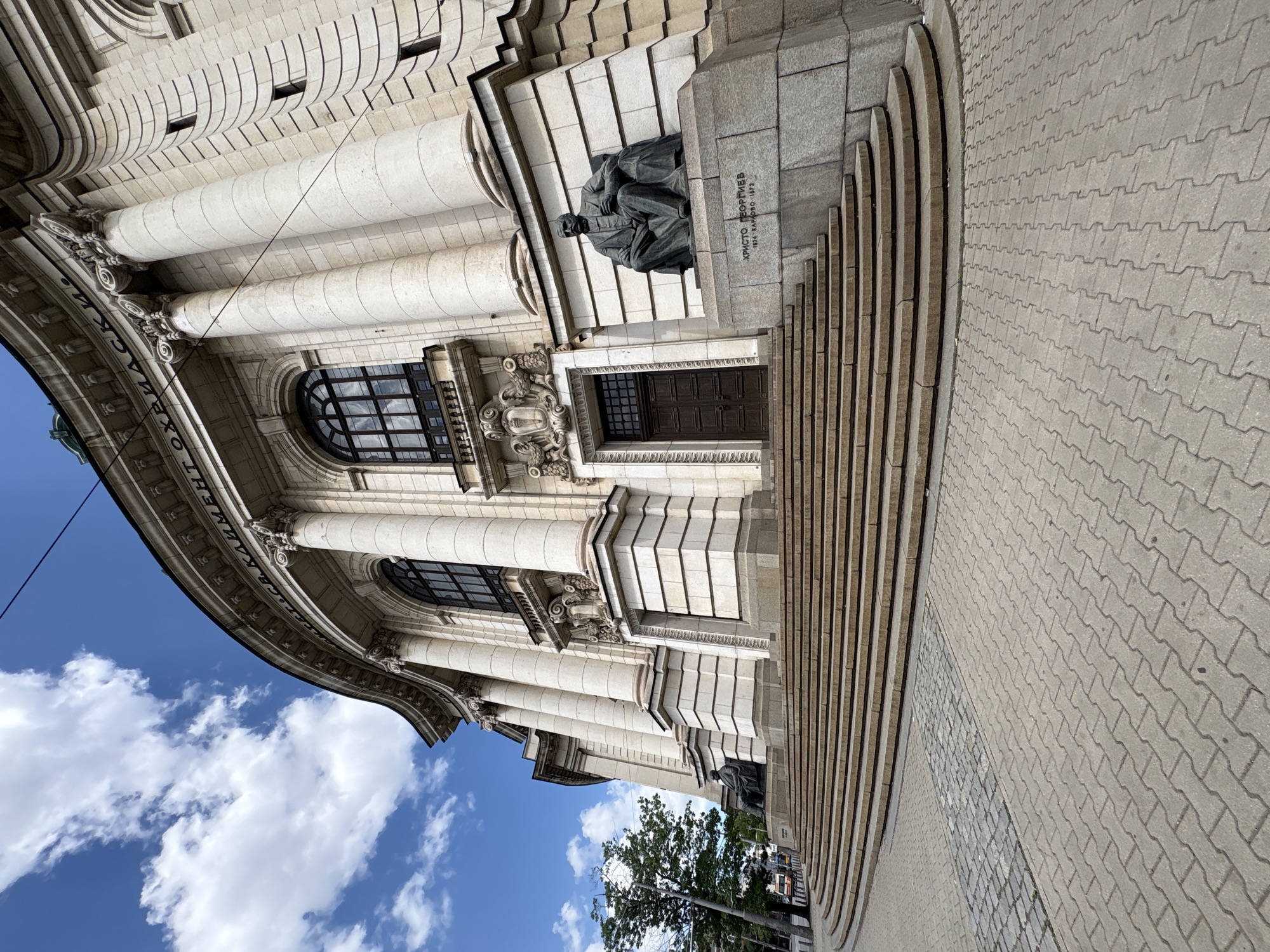 This image depicts the main facade of Sofia University "St. Kliment Ohridski" in Sofia, Bulgaria, viewed from a low angle on a sunny day. The dominant feature is the grand, curved building constructed from light-colored stone, exhibiting neoclassical architectural elements.
The building's upper section features a prominent inscription in Cyrillic, partially visible, which reads "УМЕНТ СВ. КЛИМЕНТ ОХРИДСКИ" (likely part of "Софийски университет СВ. КЛИМЕНТ ОХРИДСКИ"). A series of large fluted columns with ornate capitals supports the structure, interspersed with tall arched windows framed by dark grilles. Decorative friezes and pediments are visible above the windows and entrances. Two large, dark brown double doors are set into the lower part of the facade.
A wide, semi-circular staircase with multiple shallow steps leads up to the building's base. On the right side of the stairs, a bronze statue of a seated man, identified by a stone inscription as "ХРИСТО ГЕОРГИЕВ 1824 КАЛОФЕР-1872," rests on a stone pedestal. Another similar, but less distinct, statue is visible to the left, further down the building's facade.
The ground in the foreground is paved with interlocking concrete pavers, transitioning to a strip of cobblestones closer to the stairs. The sky is bright blue with scattered white cumulus clouds. To the far left, a portion of a tree with green foliage and a street lamp are visible, suggesting an urban street setting beyond. A faint glimpse of a road and a vehicle can be seen in the distance on the left.