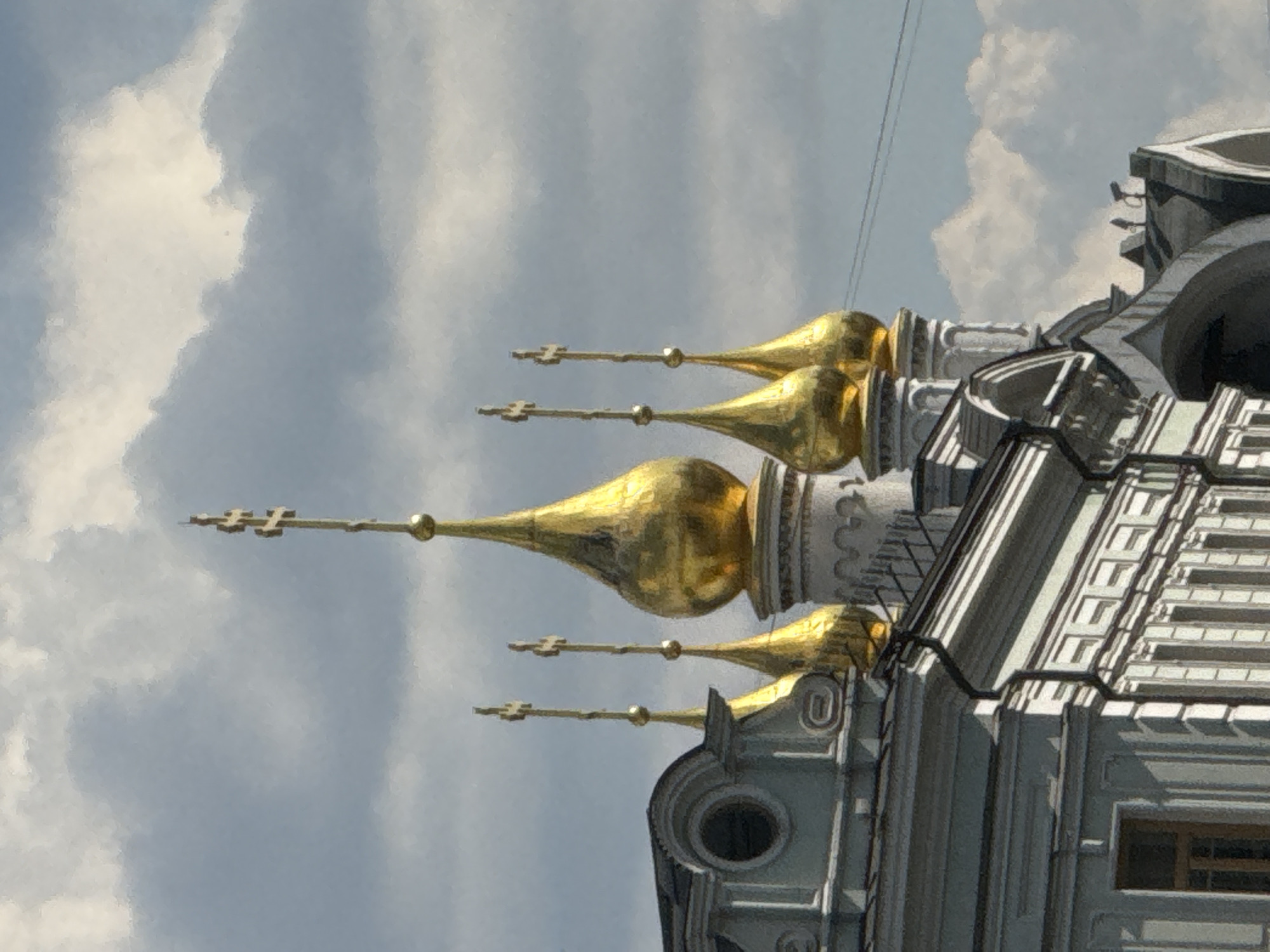 The image displays an upward view of the upper section of a majestic religious edifice, characteristic of a Russian Orthodox church or cathedral, set against a bright, partly cloudy sky. The main subjects are four prominent, highly reflective golden onion domes, each topped with an ornate three-barred Orthodox cross. These domes gleam under the sunlight, suggesting a clear day.
The building beneath the domes is constructed from light grey or white stone, featuring intricate architectural details such as layered cornices, decorative moldings, and possibly pilasters. A round window and a rectangular window with dark frames are visible on the building's facade towards the right side. Part of a patterned or tiled roof structure is also visible.
The sky in the background is a mix of soft blue and white, with scattered cumulus clouds. Thin, dark wires or cables stretch diagonally across the upper right portion of the frame. The low-angle perspective emphasizes the height and grandeur of the structure. The setting is Moscow, Russia, as per the provided context, aligning with the distinctive architectural style. There are no visible people, specific activities, or discernible text in the image.