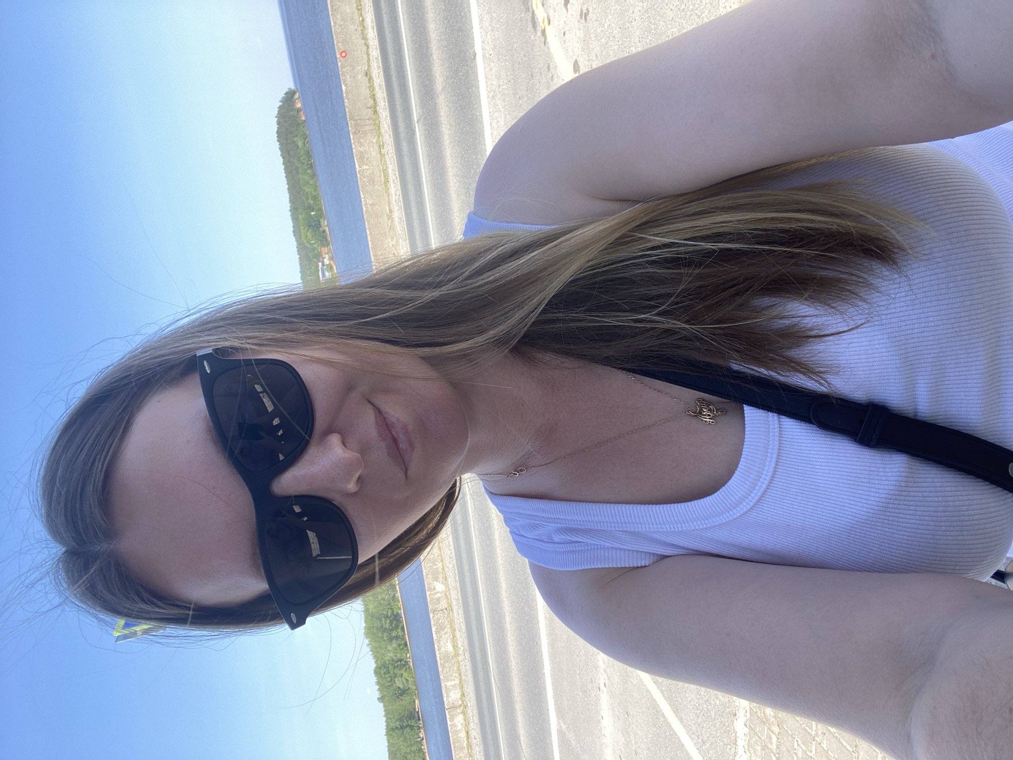 A selfie, in portrait orientation, depicts a woman wearing sunglasses and a white tank top standing outside in daylight. She has light skin and light brown hair with blonde highlights. A black strap is visible over her shoulder. A gold necklace with a charm is around her neck. In the blurred background, there's a body of water with trees along the shoreline and a bridge. The location appears to be a bridge, likely located in Noviy, Russia. The woman is holding the camera at arm's length to capture the selfie.
