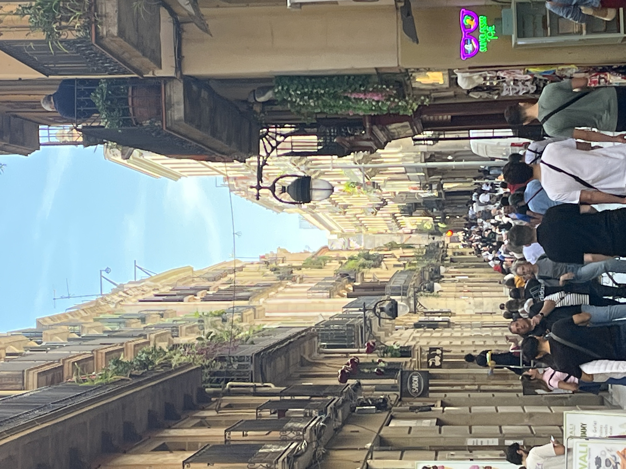 The image captures a vibrant, narrow street scene in Barcelona, Spain, under a clear blue daytime sky. The street is flanked by multi-story buildings characterized by traditional architecture, featuring numerous windows and balconies, many of which are adorned with potted plants and hanging foliage.
A significant crowd of people fills the street, suggesting a busy commercial or tourist area. Individuals are seen walking, strolling, and observing their surroundings. On a balcony to the left, a person is visible leaning out, observing the street below. The street is illuminated by ornate, black iron street lamps mounted on the building facades.
Several commercial signs are visible, including a prominent purple and green neon sign shaped like sunglasses, which clearly displays the word "Sunglasses" alongside the number "36". Further along the street, a circular shop sign reads "SABON". Another white sign with green lettering is partially visible, showing words like "ALI JIMMY" and "WALF". The bright daylight and bustling activity indicate a lively atmosphere.