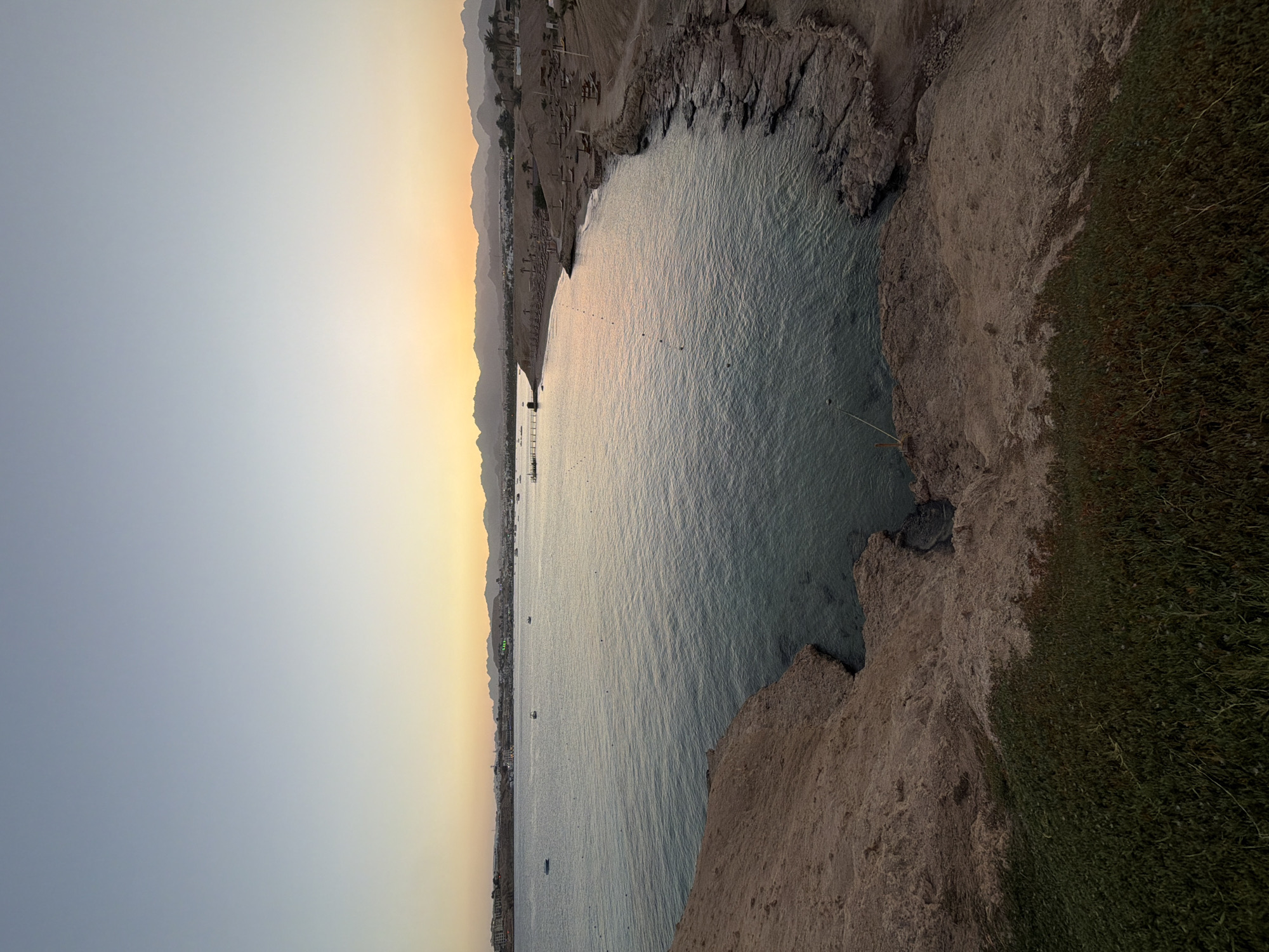This image, set in Sharm El Sheikh, Egypt, captures a tranquil coastal scene, likely during sunset or sunrise. The perspective is from an elevated vantage point overlooking a calm bay or inlet.
The sky, dominating the upper portion, transitions from a clear, pale grey-blue overhead to a warm golden-orange closer to the horizon, where distant mountains are silhouetted.
The central part of the image is occupied by the sea, which appears calm with gentle ripples, reflecting the soft hues of the sky. Several small, dark objects, possibly buoys or boats, are dotted across the water's surface.
Along the distant shore to the left, a built-up area with numerous buildings and structures, indicative of a town or resort, can be seen. This coastline appears to include a beach area. Behind this settlement, a range of mountains stretches across the horizon.
In the foreground and to the right, rugged, light brown rocky terrain dominates, sloping down towards the water. Portions of these cliffs are covered with patches of dark green, coarse vegetation, possibly grass or low shrubs.
The overall atmosphere is peaceful and serene. No people or specific activities are distinctly visible. There is no visible text in the image.