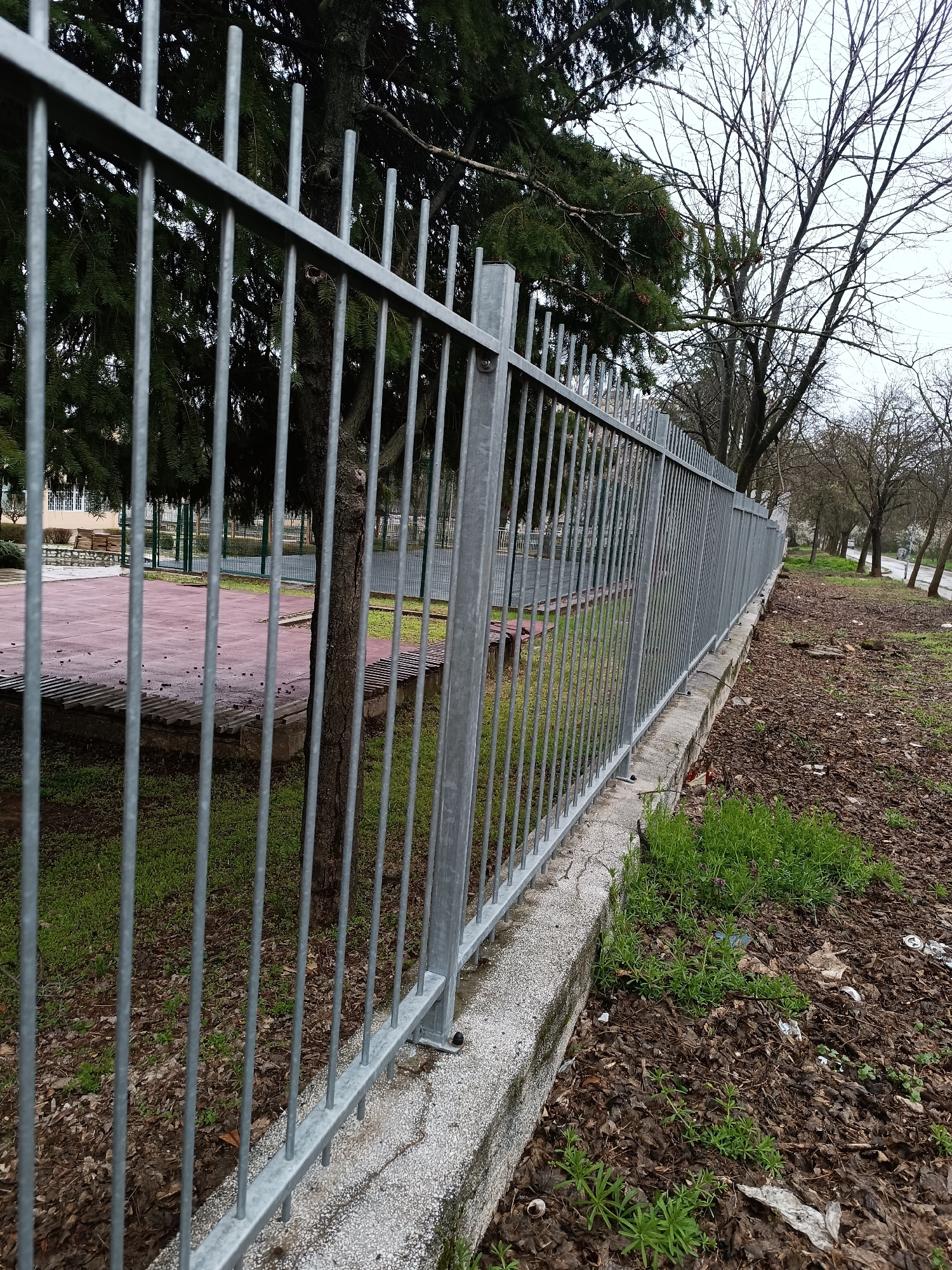 The image depicts a gray metal fence running alongside a sidewalk or path, possibly in a park or public space. The fence is set upon a low concrete base. On the left side of the fence, there is a red paved area and some vegetation. Behind the fence are various trees, some with green foliage and others bare. The ground on the right side of the fence is covered in leaves and some sparse green vegetation. The weather appears overcast or gray. The image location is Pleven, Bulgaria.