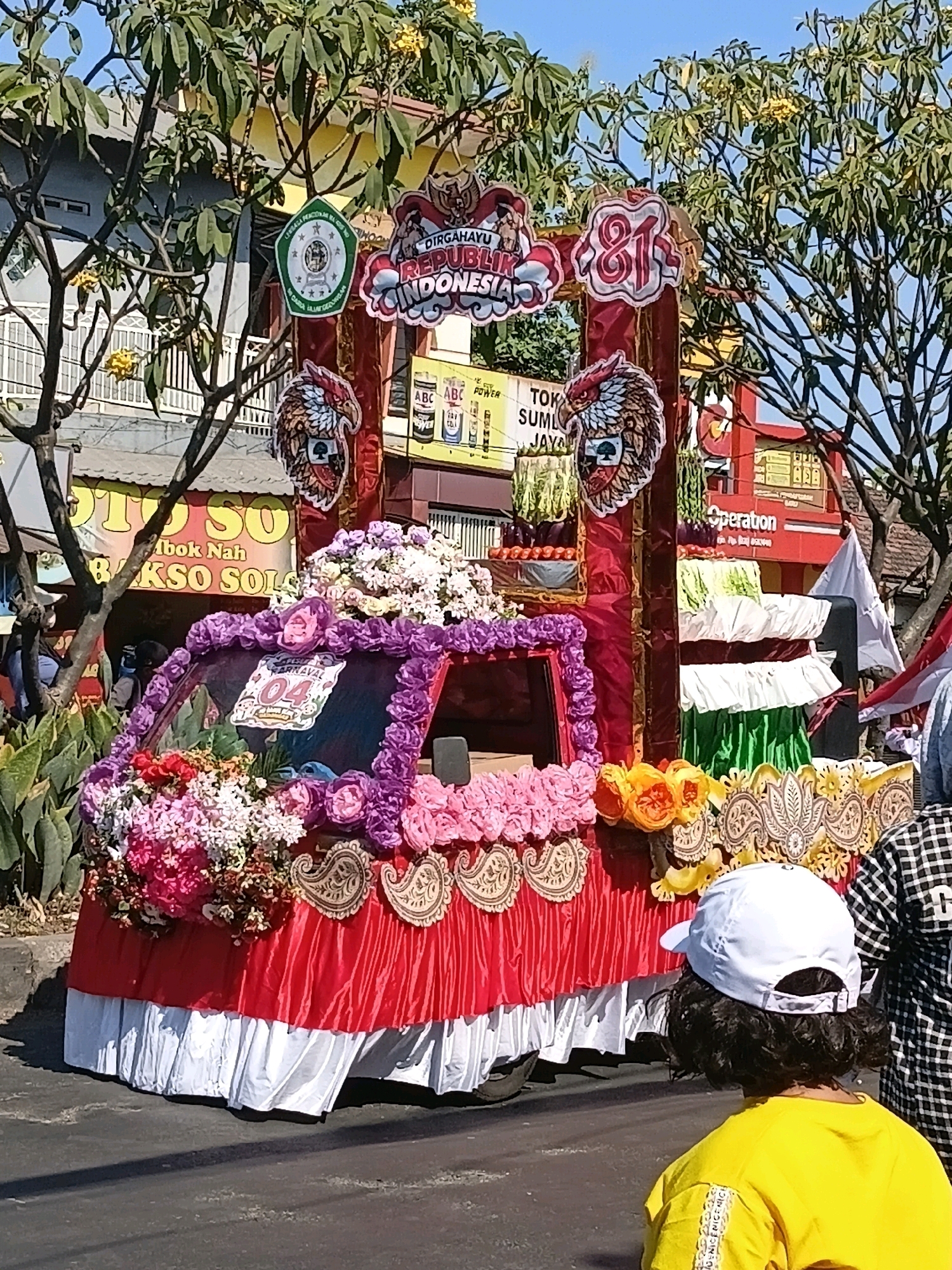 The image captures a vibrant parade float, adorned with numerous flowers in shades of pink, purple, and yellow, as well as decorative fabric. The float is decorated with banners and emblems, one of which reads "DIRGAHAYU REPUBLIK INDONESIA" and another features the number "81". A truck forms the base of the float, its windshield framed by floral arrangements. In the foreground, a person wearing a yellow shirt and a white cap is looking towards the float. In the background, buildings and trees are visible, suggesting an outdoor urban setting. The overall scene appears to be a celebratory event, possibly an Independence Day parade, taking place in Wadungasri, Indonesia.