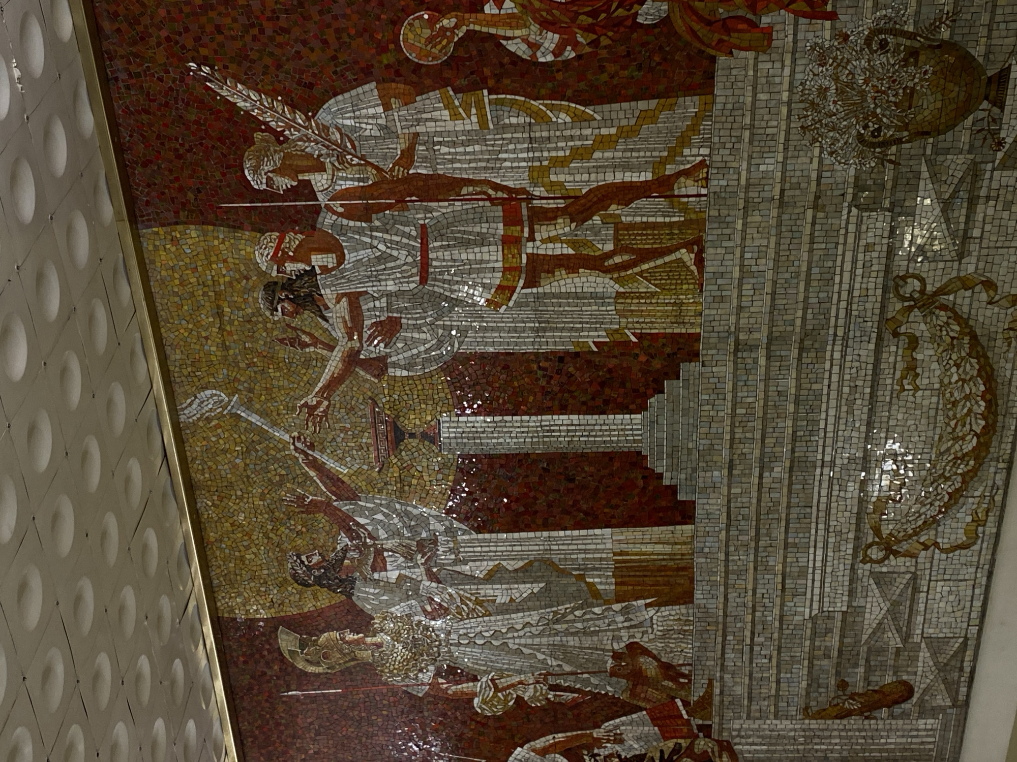 The image shows a mosaic in the Avtovo metro station in Saint Petersburg, Russia. The mosaic depicts figures in classical-style clothing, possibly representing ancient warriors or heroes. The scene includes human figures in various poses: some standing, some falling, and one on their knees, with spears. The mosaic is composed of small tiles of different colors, mainly shades of gold, brown, red, and white. To the right of the mosaic is a gray tiled pattern, and a decorative vase and ribbon motif can also be seen. The mosaic is attached to a wall that has a pattern with concave circles on it.