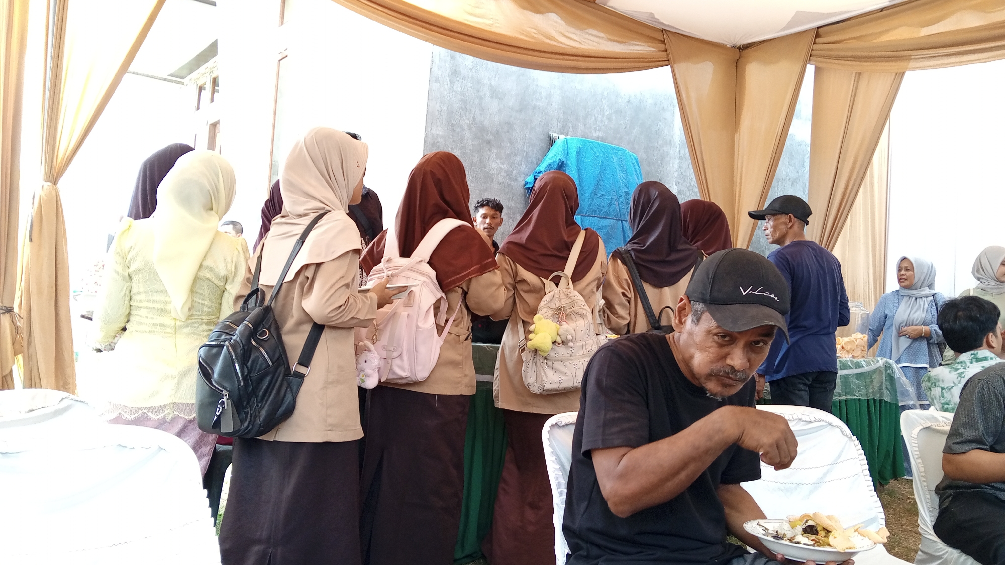 The photograph captures a gathering of people under a makeshift tent structure, likely for an event, in Payakumbuh, Indonesia. The scene is somewhat crowded, with individuals of various ages present, predominantly dressed in modest attire. Several women are wearing hijabs, common in Indonesia. In the foreground, a man with a mustache wearing a black baseball cap and a black t-shirt is holding a plate of food and looking towards the camera. To his right, another man in a dark blue shirt and a black cap is visible. Behind them, a group of young women, some in matching beige uniforms and hijabs, are standing, with at least two carrying backpacks. One backpack is adorned with a yellow plush toy. Further back, other individuals can be seen mingling, and a table with snacks is present. The environment appears to be outdoors, with a grey wall visible in the background, and beige drapes creating the tent enclosure. The overall impression is of a social gathering or celebration taking place in a community setting.