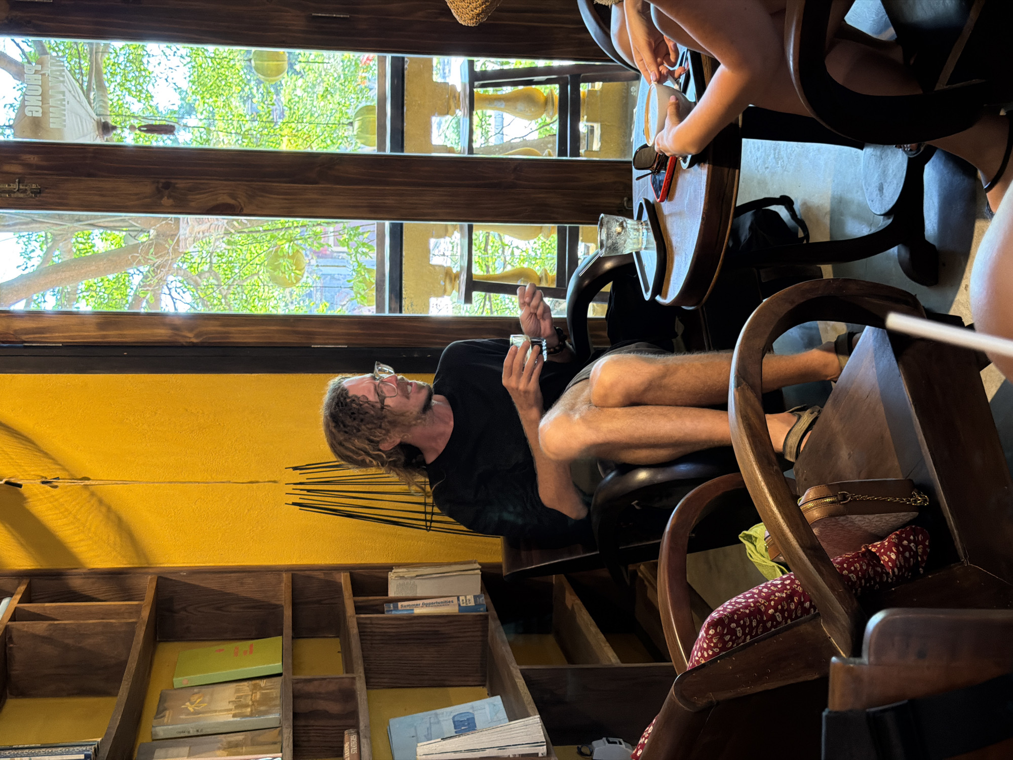 The image depicts a scene inside a cafe in Hội An, Vietnam, characterized by a warm yellow wall and wooden furnishings. A man with curly hair and glasses, wearing a black t-shirt and shorts, is seated in a dark wooden chair, holding a glass. He appears to be engaged in conversation. In the foreground, a round wooden table holds drinks, including a coffee cup, and a smartphone. Two individuals, partially visible, are seated at the table, with one holding the coffee cup. Another person's legs and feet are visible in the lower right corner of the image, indicating at least three people are present. Behind the man, a tall wooden bookshelf is filled with books. The cafe's ambiance is enhanced by natural light filtering through wooden-framed windows, revealing lush green foliage outside.