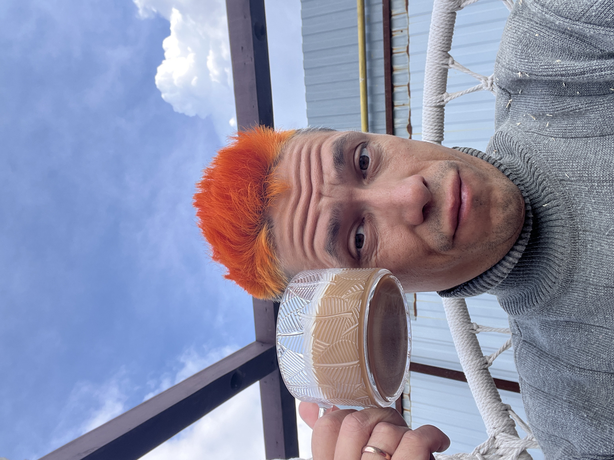 A man with short, orange hair holds a glass mug filled with a tan-colored beverage. He wears a gray turtleneck sweater. A wedding band is visible on his finger. The background includes a cloudy sky, a metal structure, and a rope chair. Based on available information, it is difficult to confirm the location specifically as Chaikovsky, Russia.