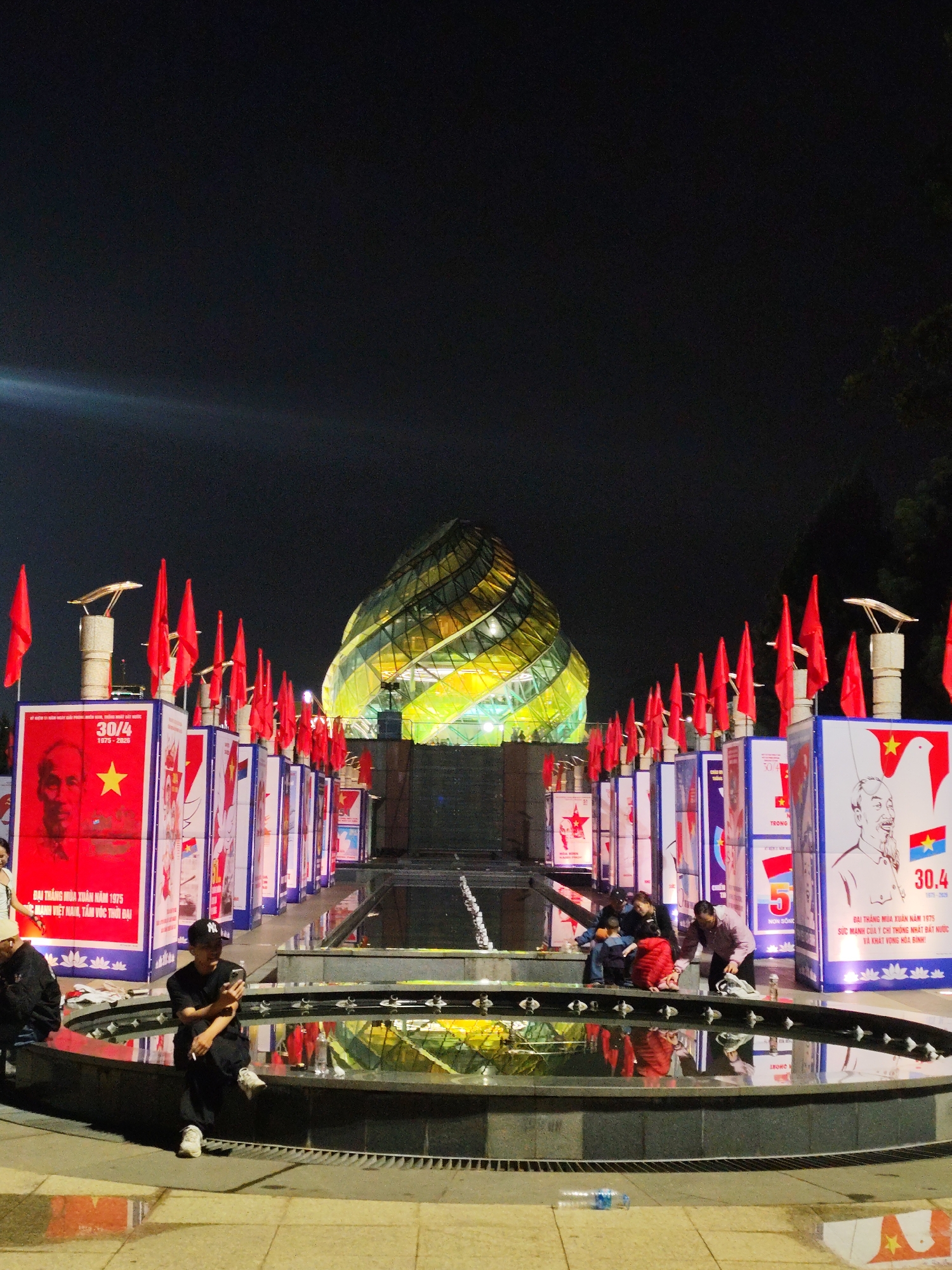 This is a nighttime image of a public space in Da Lat, Vietnam. In the background is a large, dome-shaped structure illuminated in hues of yellow, orange, and green. It appears to be constructed of glass or a similar transparent material. Arranged in a row leading towards the structure are rectangular stands, each topped with a red flag. These stands are adorned with imagery related to Vietnamese political themes, including portraits of historical figures and dates like "30/4," likely referring to the Reunification Day of Vietnam.
In the foreground, there is a circular fountain or reflecting pool. Several people are sitting around the edge of the fountain, some are seated, others are leaning over looking into the water. There is a male on the left sitting on the rim of the fountain who is looking at his phone. The scene is well-lit.