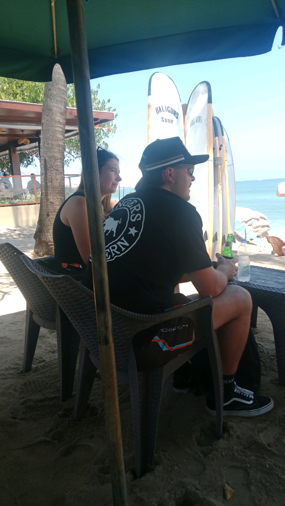 In Kuta, Indonesia, two individuals are seated at a table on a sandy beach. The person on the left, a woman with long brown hair, is wearing a black tank top. The person on the right, a man wearing a black baseball cap and sunglasses, has a black t-shirt with a circular logo featuring a bull and text that appears to read "BALIGUNS SURF" and "WESTERN". He is holding a green bottle and a plastic water bottle. In the background, several surfboards are propped up. The scene is shaded by a large green umbrella. The environment suggests a casual beachside setting, likely a cafe or bar, with the ocean visible in the distance.