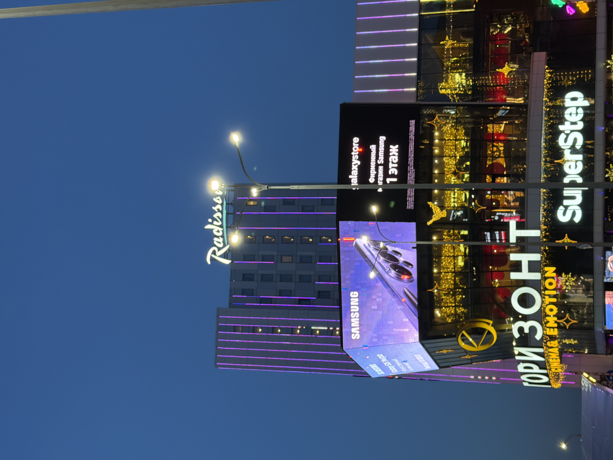 Here's a description of the image:
The image is a nighttime shot of buildings in Rostov-on-Don, Russia, featuring prominent advertisements.
A building with the "Radisson" name in illuminated letters is visible, along with a Samsung advertisement. The Samsung ad shows a smartphone, presumably a Galaxy model. "Galaxystore" is also advertised as a "flagship Samsung store" on the 1st floor.
To the right, other signs are visible, including "Горизонт" (Horizon), advertising "CINEMA & EMOTION" as well as "SuperStep." The buildings are decorated with lights and signs, indicating a commercial or entertainment area. The sky is a deep blue, suggesting it's dusk or early evening. Streetlights are present.
It's a street scene in the city of Rostov-on-Don, Russia, with buildings illuminated by lights and advertising signs.