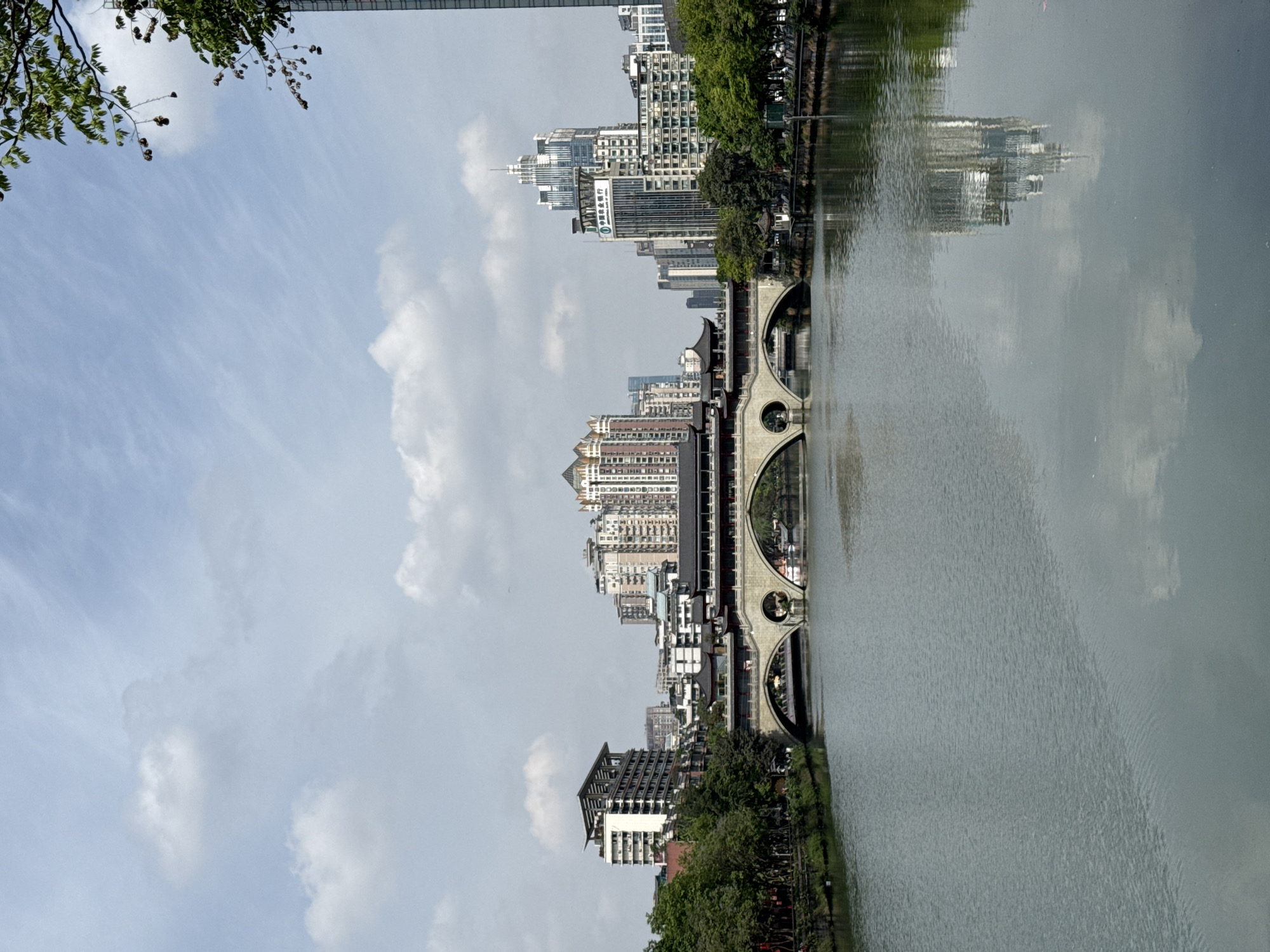 The image captures a picturesque cityscape in Jinjiang District, China, on a bright, partly cloudy day. The sky is a soft blue with a scattering of white clouds, ranging from wispy cirrus to more defined cumulus formations. In the upper left corner, green tree branches with small, dark seed pods extend into the frame, partially obscuring a distant bridge or structure.
A calm river dominates the foreground and middle ground, acting as a reflective surface that mirrors the sky and the urban landscape. Spanning the river is an elegant multi-arched bridge, characterized by its distinctive circular cutouts above each arch and a flat top likely serving as a pedestrian walkway.
Along the far bank of the river, a diverse array of buildings rises. These include modern high-rise glass and concrete skyscrapers alongside structures featuring traditional Chinese architectural elements, such as curved roofs. Prominently visible on a tall, light-colored modern building, the vertical Chinese characters "中国民生银行" (China Minsheng Bank) are discernible. Lush green trees and foliage line the riverbanks, providing a natural contrast to the dense urban environment. The scene depicts a serene daytime view of a bustling city integrated with natural waterways and traditional aesthetics.