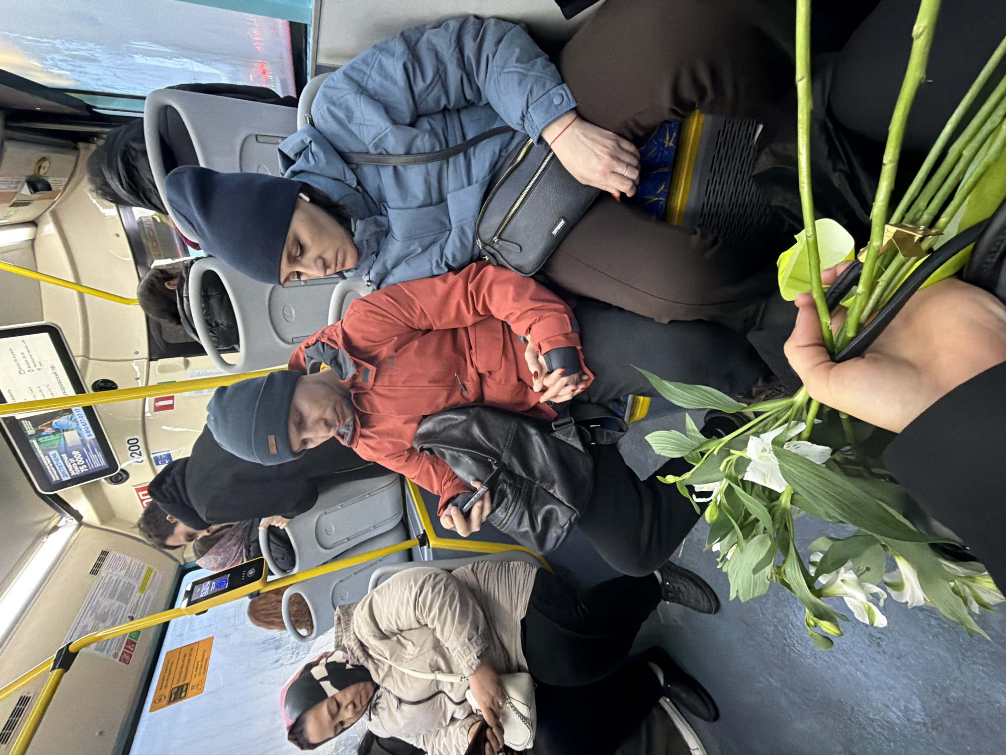 The image captures a scene inside a public bus in Saint Petersburg, Russia. Several passengers are seated, and some appear to be asleep. A woman wearing a blue puffy jacket and a dark blue beanie is resting her head on another person, who is wearing an orange jacket and a dark beanie. A handbag is draped across her lap.
Further down the aisle, another passenger, wearing a beige jacket and a patterned head covering, is also seated. In the foreground, someone is holding a bouquet of white lilies. The bus's interior is visible, including seats, handrails, and information displays, one of which has the text "8-812-677-07-17" and a salary advertisement of "75000P". The text "Уворовский пр." (Uvarovsky prospect in English) is also visible, likely indicating the location of the bus.