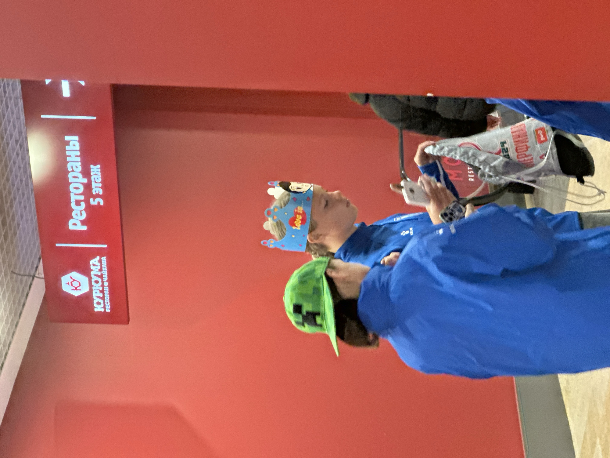 The image shows two children in a building. The child closest to the camera is wearing a green Minecraft Creeper cap and a blue jacket. The child behind them is wearing a blue birthday crown. The children are near a sign that reads "Рестораны 5 этаж" (Restaurants 5th floor) and "ЮРКУМА РЕСТОРАН ЧАЙХАНА" (KURKUMA RESTAURANT CHAYHANA). The image was taken in Nizhny Novgorod, Russia.