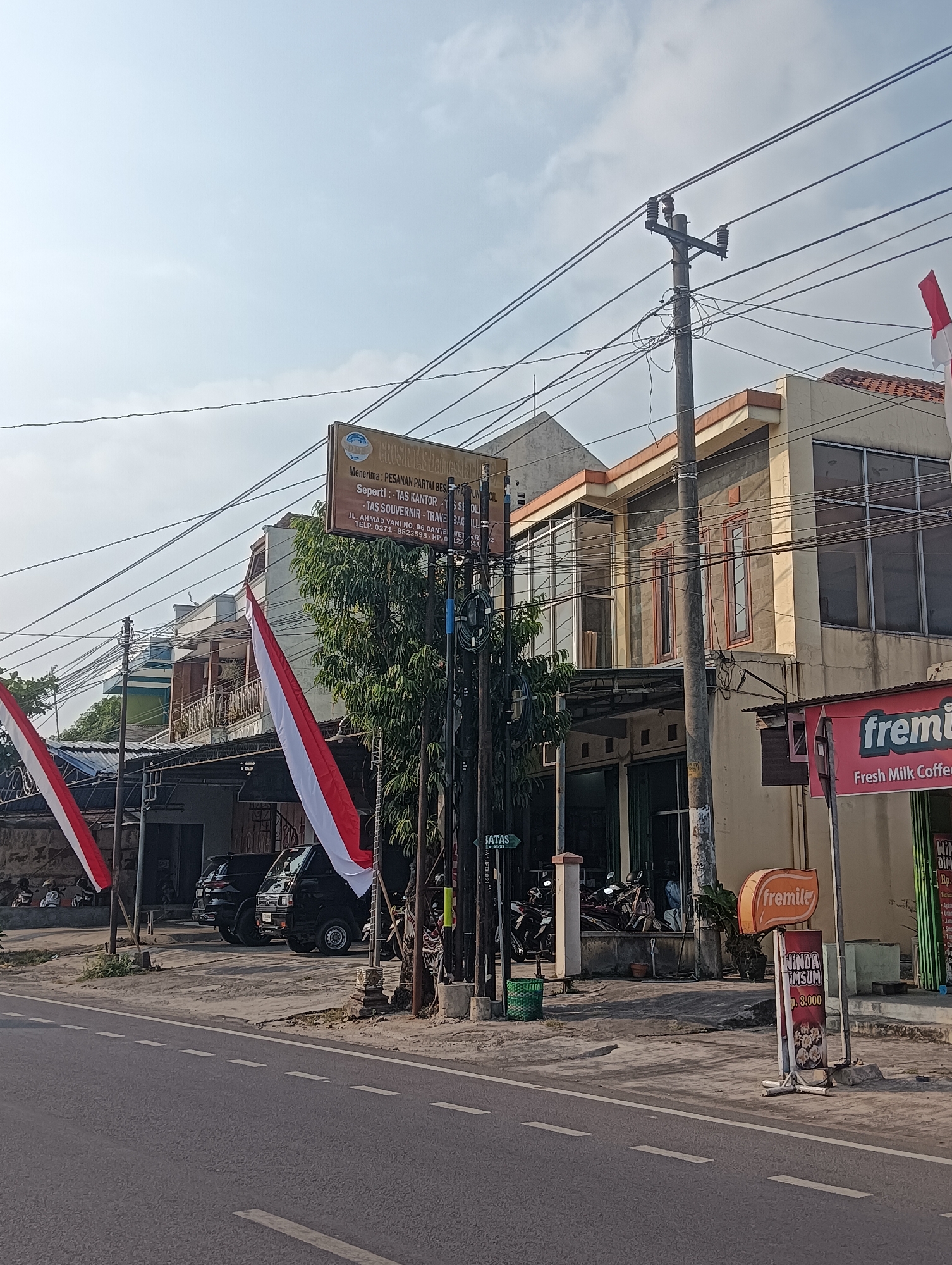 The image shows a street scene in Sragen, Indonesia. On the right side of the image is a building with a cream-colored facade, multiple windows, and a red awning with the text "fremila Fresh Milk Coffee" on it. To the left of this building, there is a sign with orange text reading "fremila" and another sign below it with red text that reads "MIND A TIMSUM Rp. 3.000". Further to the left, there is a row of buildings with balconies and some trees. Red and white Indonesian flags are hanging from poles in front of these buildings. Several vehicles, including two black vans and some motorcycles, are parked in front of the buildings. The street in the foreground has white dashed lines indicating traffic lanes. The sky is clear with some clouds.