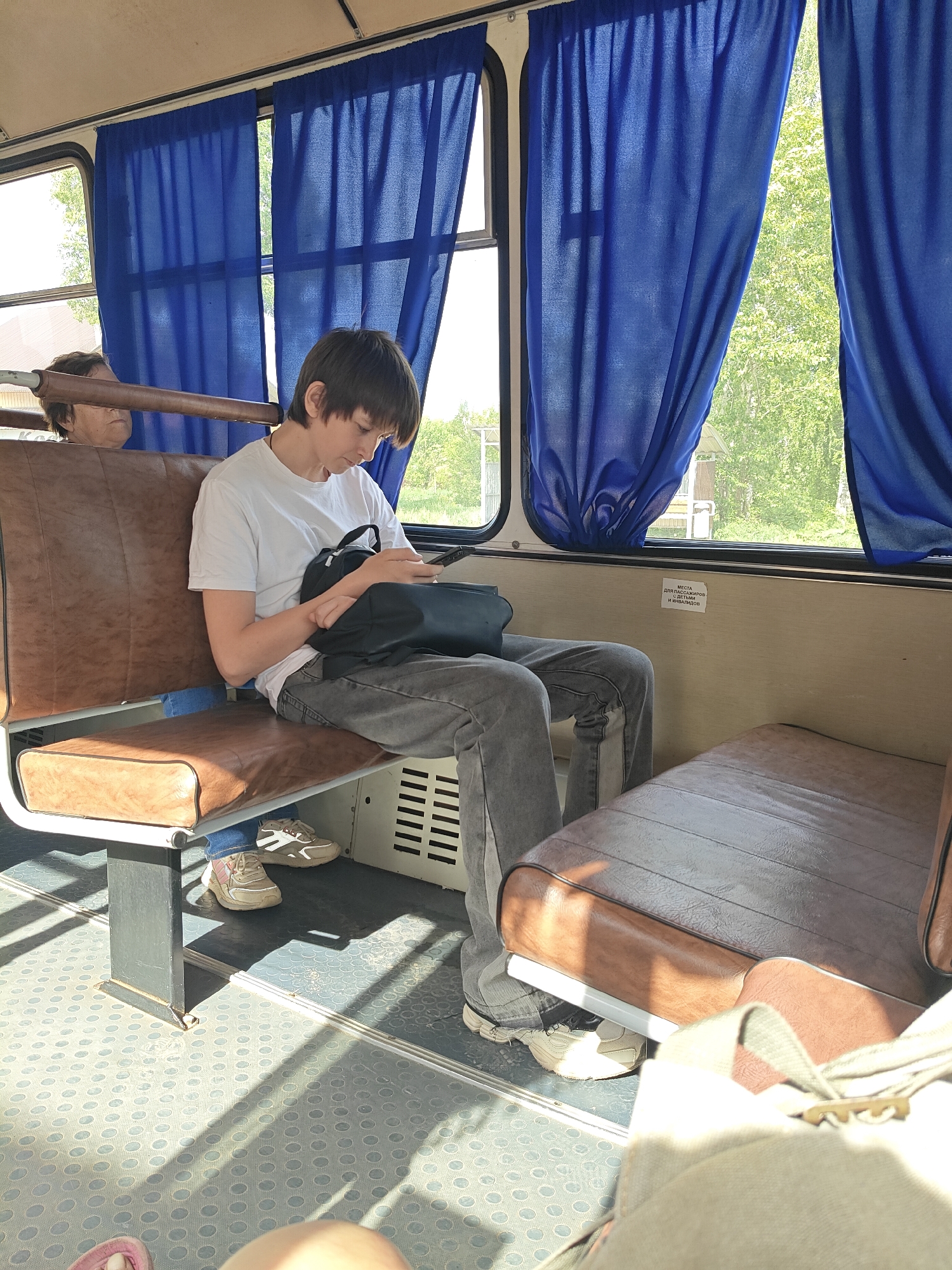 The image shows the interior of a public transportation vehicle, likely a bus, in Фоки, Russia. A young person is seated on a brown leather-like seat, occupied using a mobile phone, and carrying a black bag. The individual is wearing a white t-shirt, gray jeans, and light-colored sneakers. Through the window, a blurred outdoor scene of trees and buildings can be seen. Blue curtains hang on the window frames. Another person's head is visible behind the seat, in the top left of the image. The floor has a patterned surface. The bus interior has ventilation vents near the seats. A sign with text in Russian is attached to the wall above one of the seats.