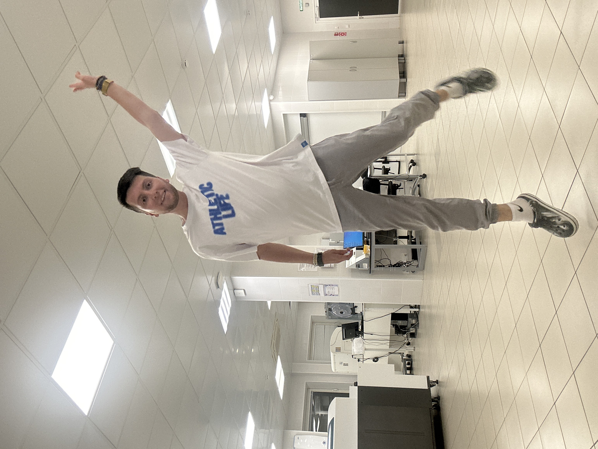 A man is jumping in a laboratory setting. He is wearing a white t-shirt with blue text that reads "ATHLETIC LIFE" and grey sweatpants. He is in mid-air, with his right arm raised and his left leg bent. The background shows a brightly lit laboratory with tiled floors, white walls, and various scientific equipment, including what appear to be centrifuges and computer monitors. There are also white cabinets and a door with a sign indicating an exit in Russian. The overall impression is of a modern, sterile research facility. The city of Moscow, Russia, is not explicitly depicted, but the presence of Russian signage on the door suggests the location could be in Moscow.
