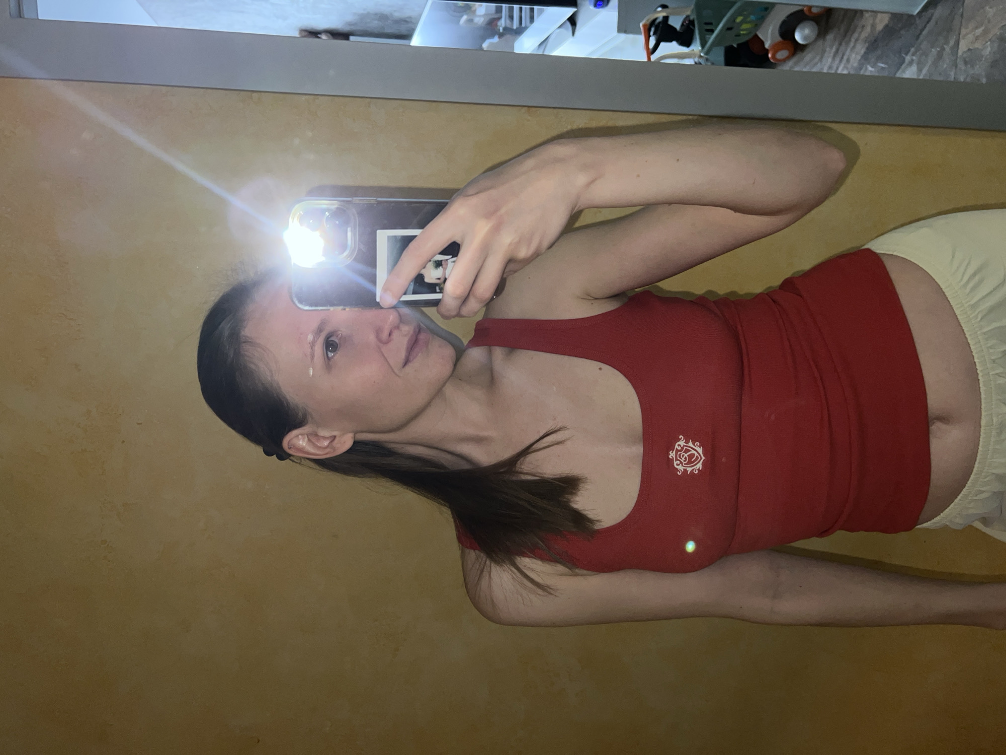 A woman with brown hair is taking a selfie in front of a mirror. She is wearing a red tank top and light yellow shorts. The mirror reflects part of a room, including what appears to be a shelf with toys. The environment and the subject's apparel do not provide information that would allow determining the location to be Khimki, Russia.