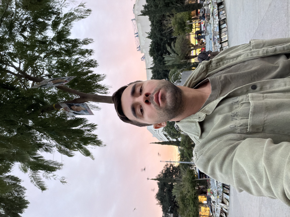 A man poses in front of a tree with photographs attached to its branches. He wears a green shirt or jacket. The background features a pale, pink-tinged sky, trees, and a structure in the distance. A vendor's display is visible in the right background. The image is rotated 90 degrees counter-clockwise.
