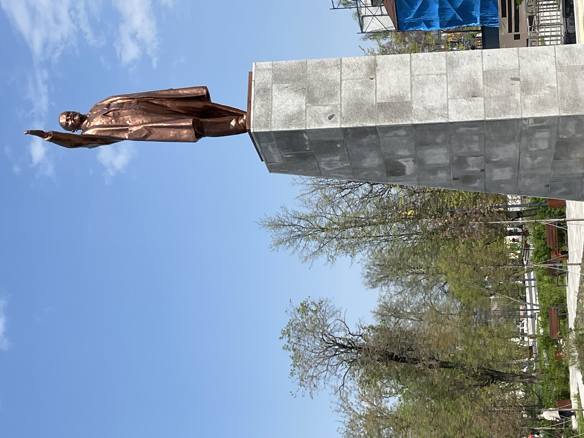 The image shows a statue of Vladimir Lenin in Noginsk, Russia. The bronze statue depicts Lenin in a dynamic pose, arm outstretched, atop a tall, rectangular, gray stone pedestal. The background features a clear blue sky, trees with budding leaves, and a partially visible building with blue scaffolding. The statue is oriented horizontally in the frame.