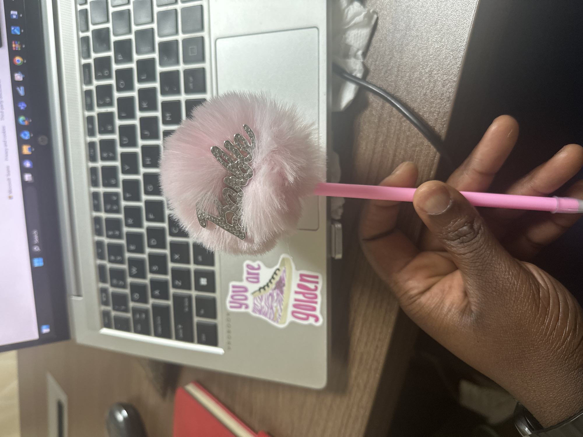 The image shows a person's hand holding a pink fluffy pen with "Barbie" written on it in glitter. The pen is positioned over a laptop keyboard. To the left of the pen, on the laptop, are two stickers: one of a pink and yellow zebra-striped high-top sneaker, and another with the text "you are golden" in purple and pink letters. The laptop is open and appears to be on a wooden desk. In the background, a red notebook and a dark-colored mouse are visible. The scene does not provide any information to place it in the city of Shaba, Nigeria.