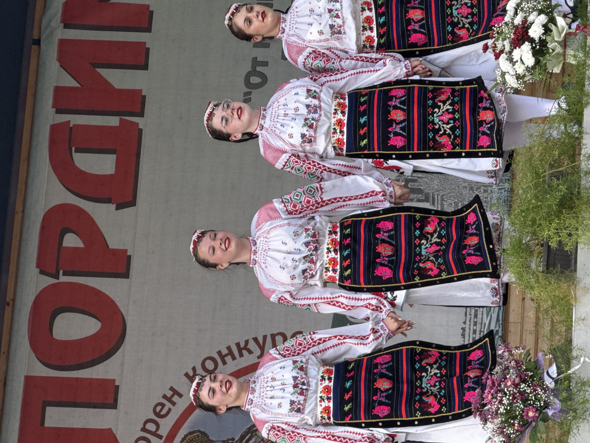 Four young women in traditional Bulgarian folk costumes are standing on a stage, holding hands. Their outfits consist of white embroidered blouses with red trim, black aprons with colorful embroidered patterns, and headbands with white ornamentation. They are smiling and appear to be performing or posing for a photograph. In the background, a large banner with red lettering is visible, partially obscured by the women. The stage is decorated with floral arrangements, including a bouquet of purple flowers on the left and white and red flowers on the right. Green foliage is also present in the foreground. The overall scene suggests a cultural event or festival taking place in Pordim, Bulgaria.