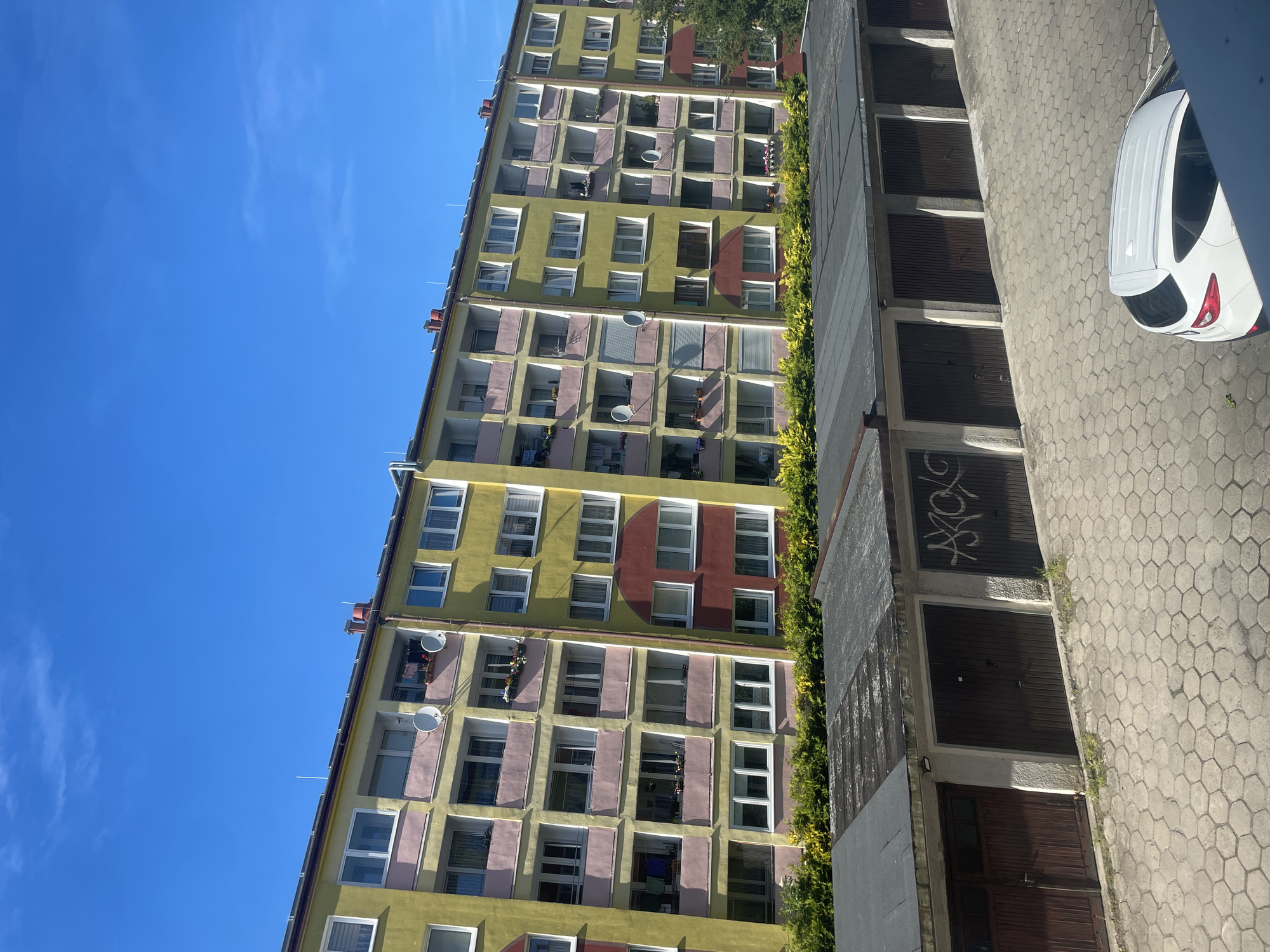 The image depicts an apartment building with a light yellow facade and pink balconies in Kołobrzeg, Poland. The building has multiple stories with numerous windows and satellite dishes visible. In front of the building, a row of garages with dark brown doors is situated. One of the garage doors is marked with graffiti. A white car is parked on a hexagonal paved area in front of the garages. Lush green bushes line the area between the apartment building and the garages. The sky above is a clear, bright blue with a few wispy clouds. The scene appears to be captured during daylight hours.