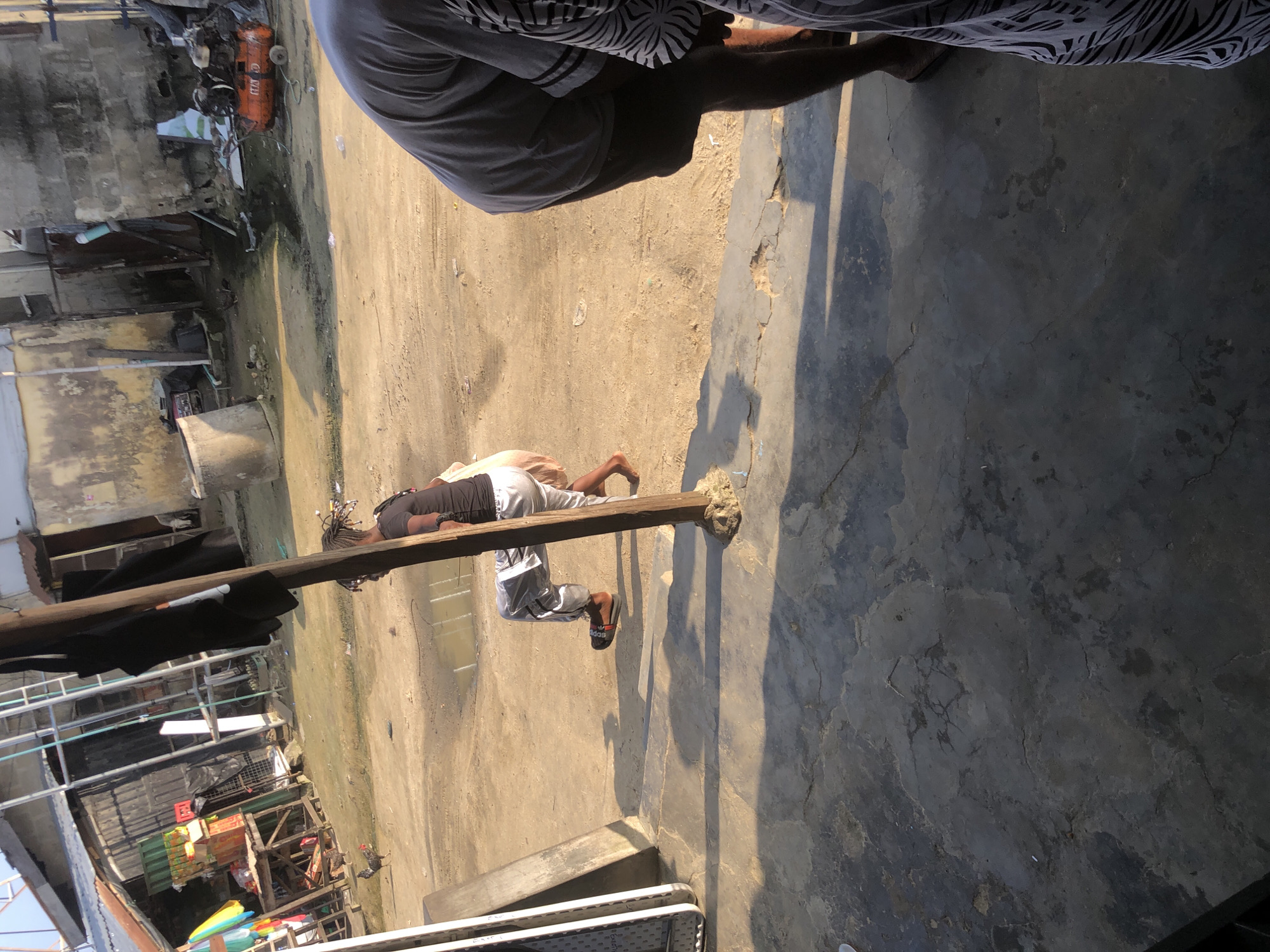 In Ado, Nigeria, a dusty, outdoor marketplace setting is depicted. A person wearing a black t-shirt and silver pants is observed in motion, leaning on a wooden post. The person appears to be moving from left to right across the frame. The ground is a mixture of sand and dirt, with a small puddle of water visible. In the background, various structures, including makeshift stalls and buildings constructed with corrugated metal and concrete blocks, line the area. A compressor is visible to the right, and several chickens are scattered throughout the scene. The foreground is dominated by a cracked concrete surface, with shadows cast by overhead structures.